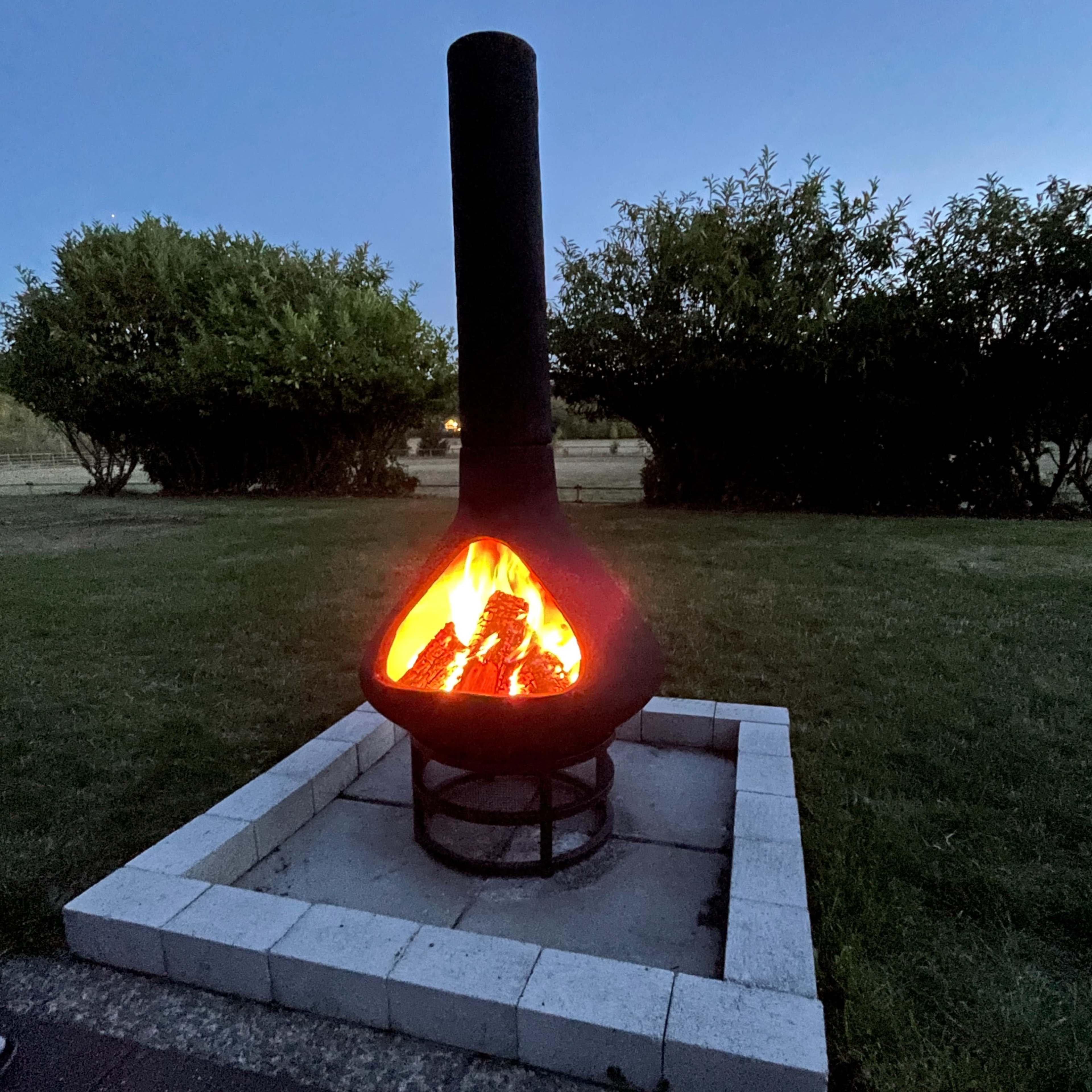 A chiminea with a fire burning inside is situated on a stone patio surrounded by greenery at dusk.
