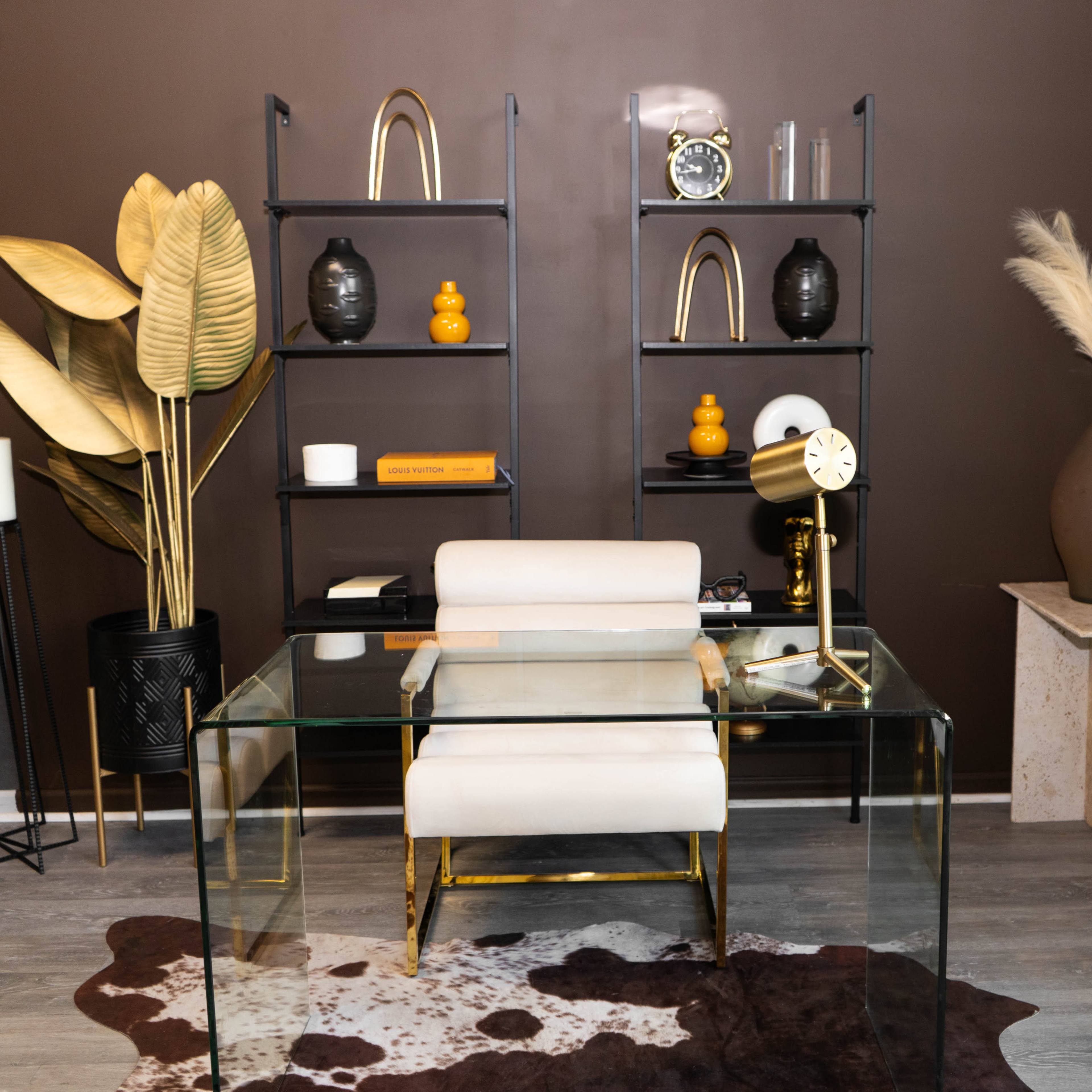 A modern office space featuring a glass desk, a white chair, and a variety of decorative items on black shelves against a brown wall.