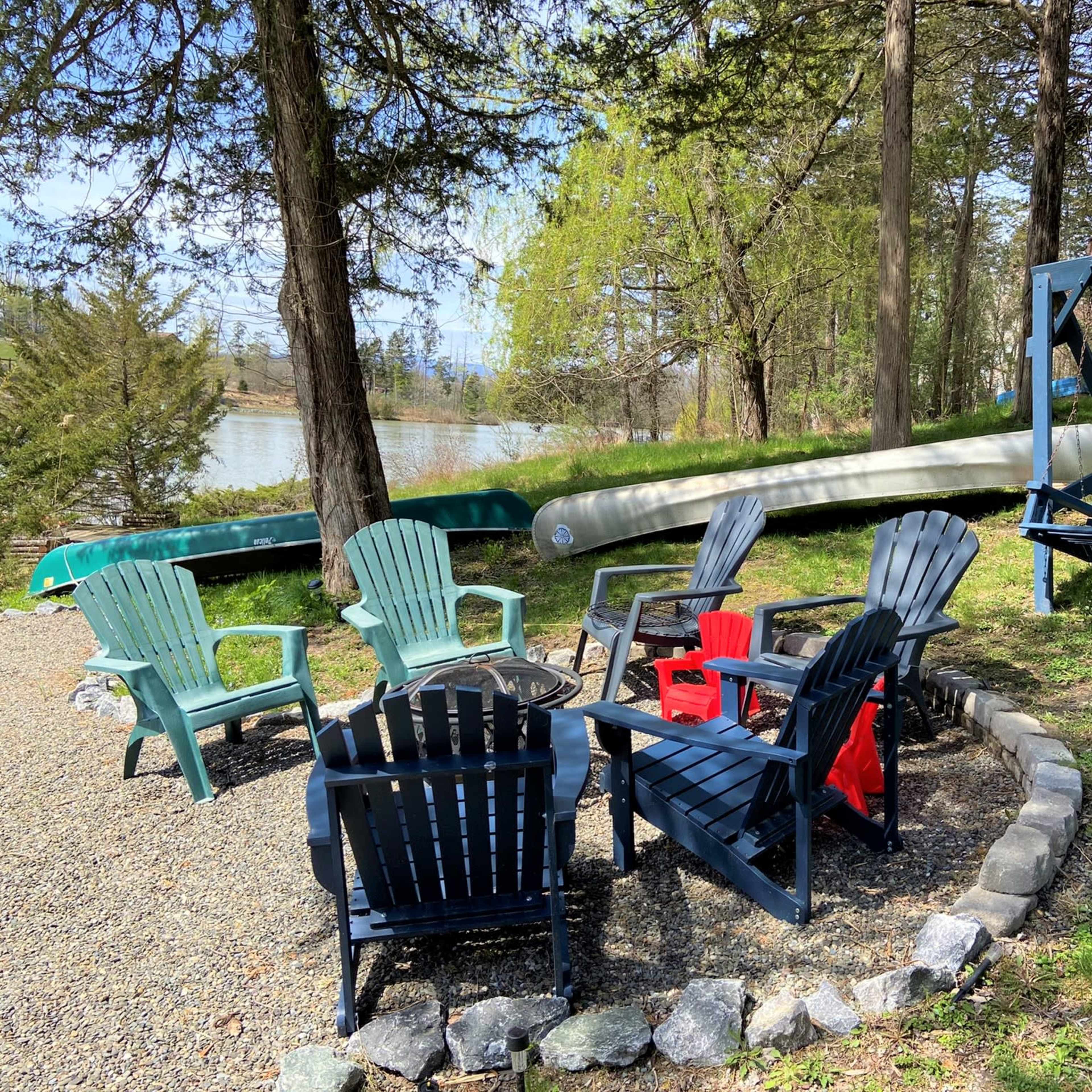 A circle of adirondack chairs surrounds a fire pit near a tranquil lake, with two canoes resting on the shore and a swing set nearby.
