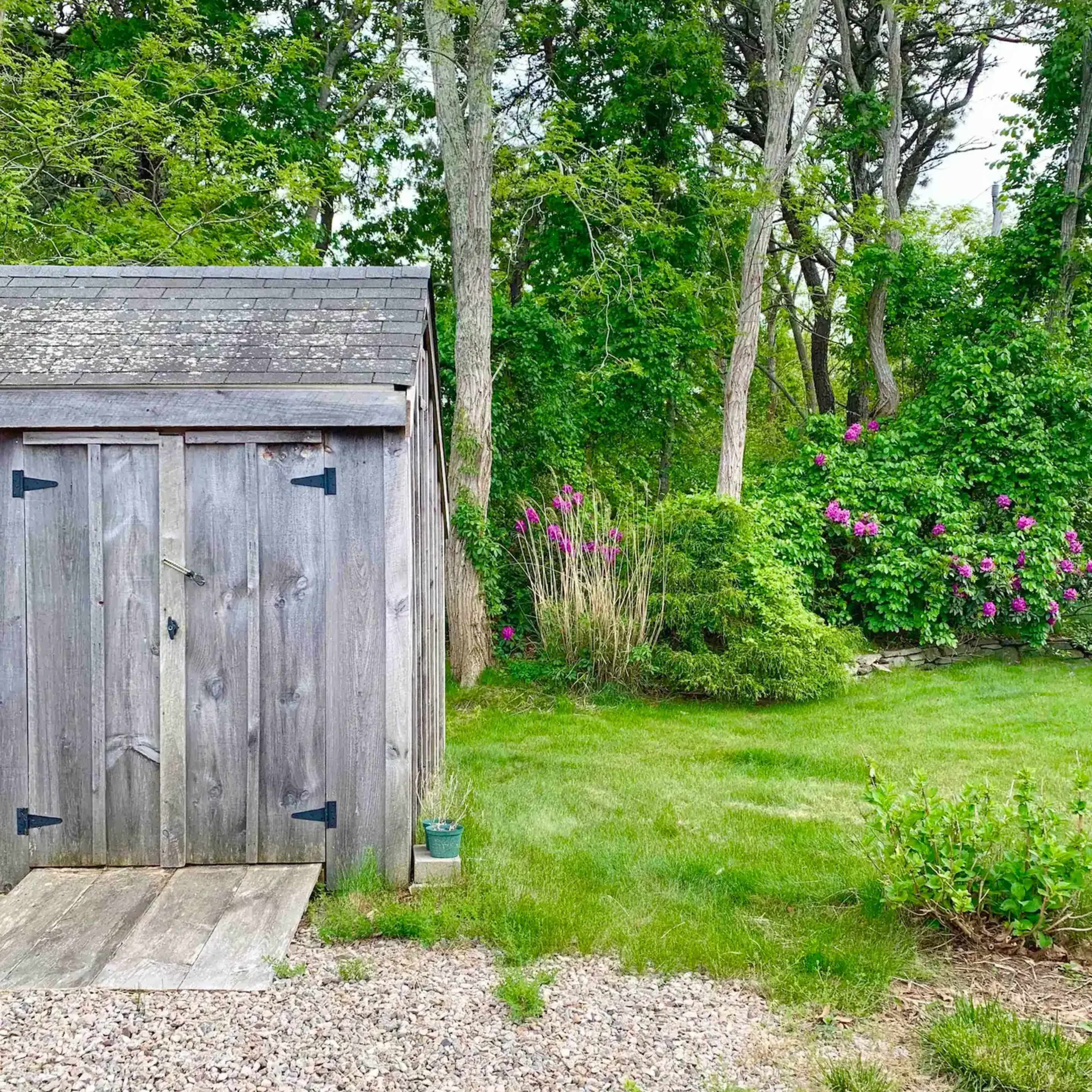A wooden shed with a sloped roof stands beside a gravel path, surrounded by lush green grass and flowering shrubs.