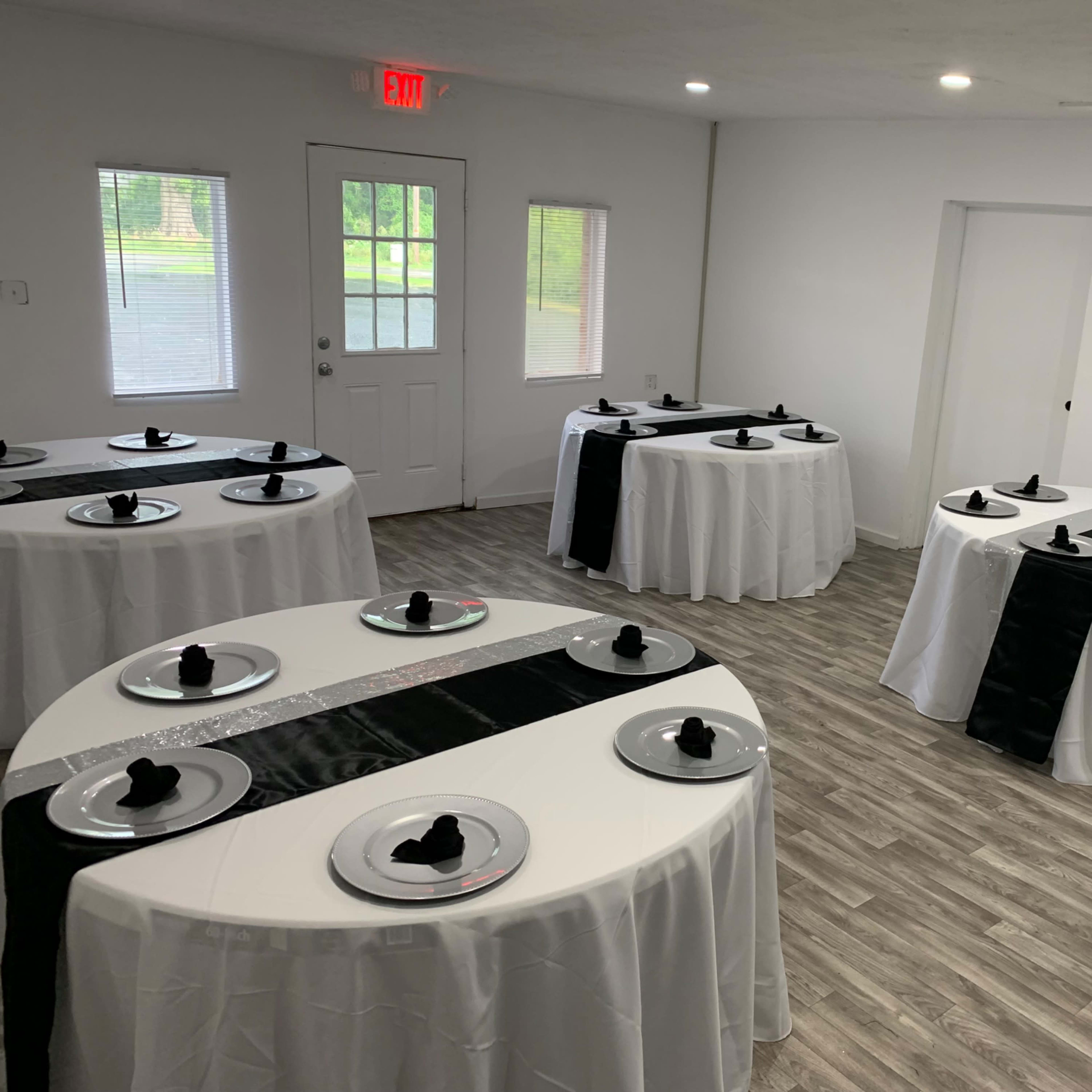 A room set up for an event with multiple round tables covered in white tablecloths, each featuring a black runner and black napkin at each place setting.