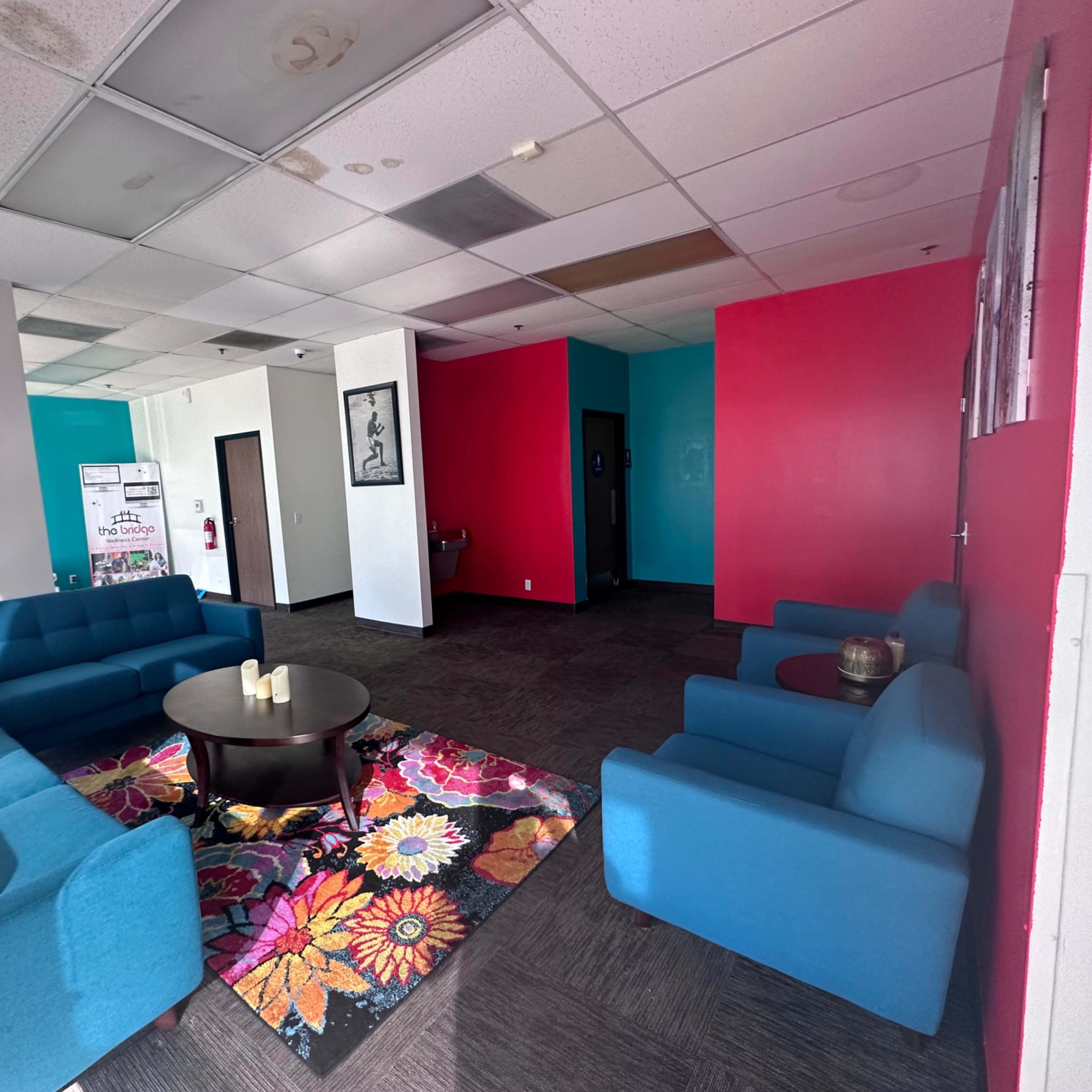 A waiting area with blue sofas, a round table, and a colorful floral rug against a backdrop of pink and white walls.