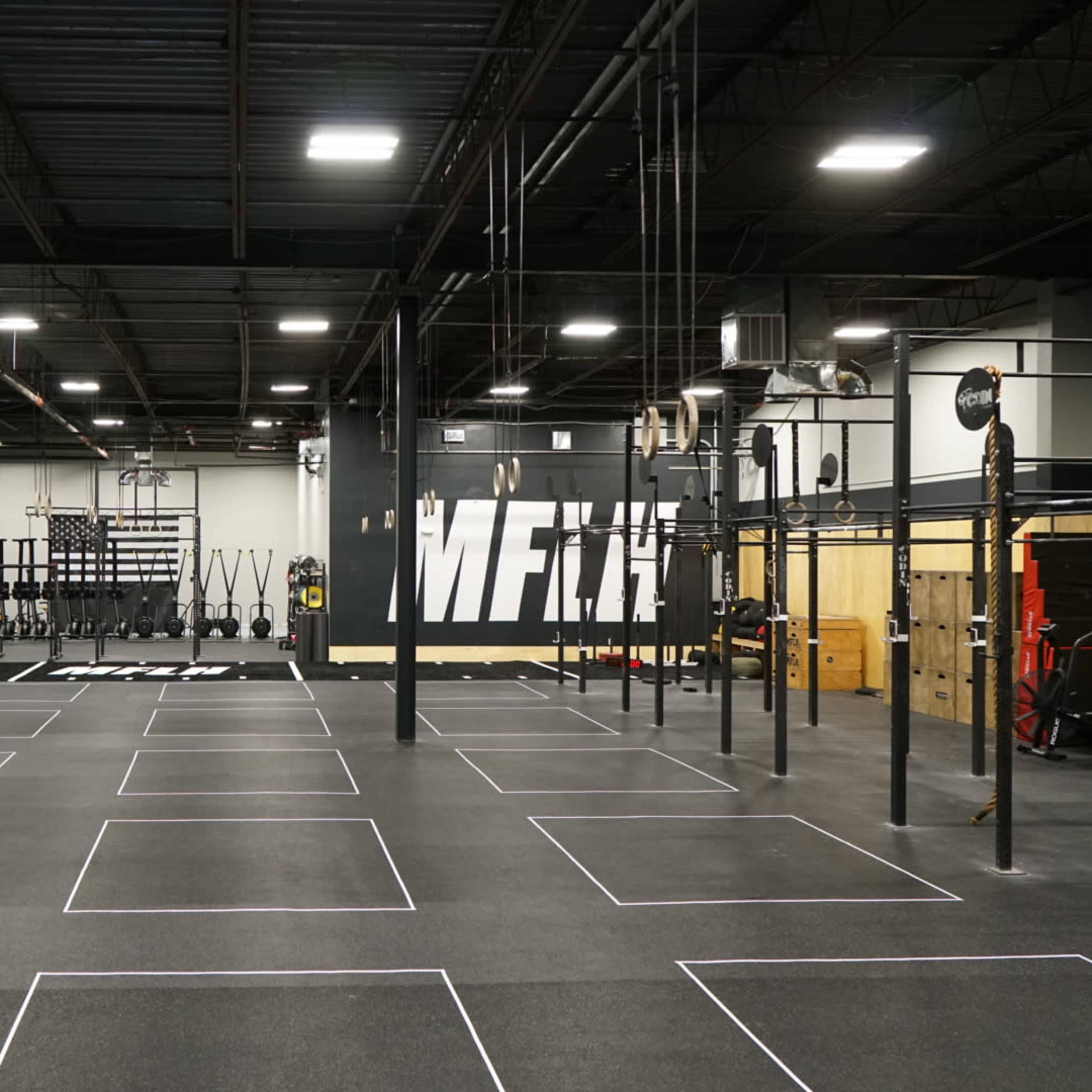 A spacious gym interior with various workout equipment, marked exercise zones, and a large wall mural that reads "MFT."