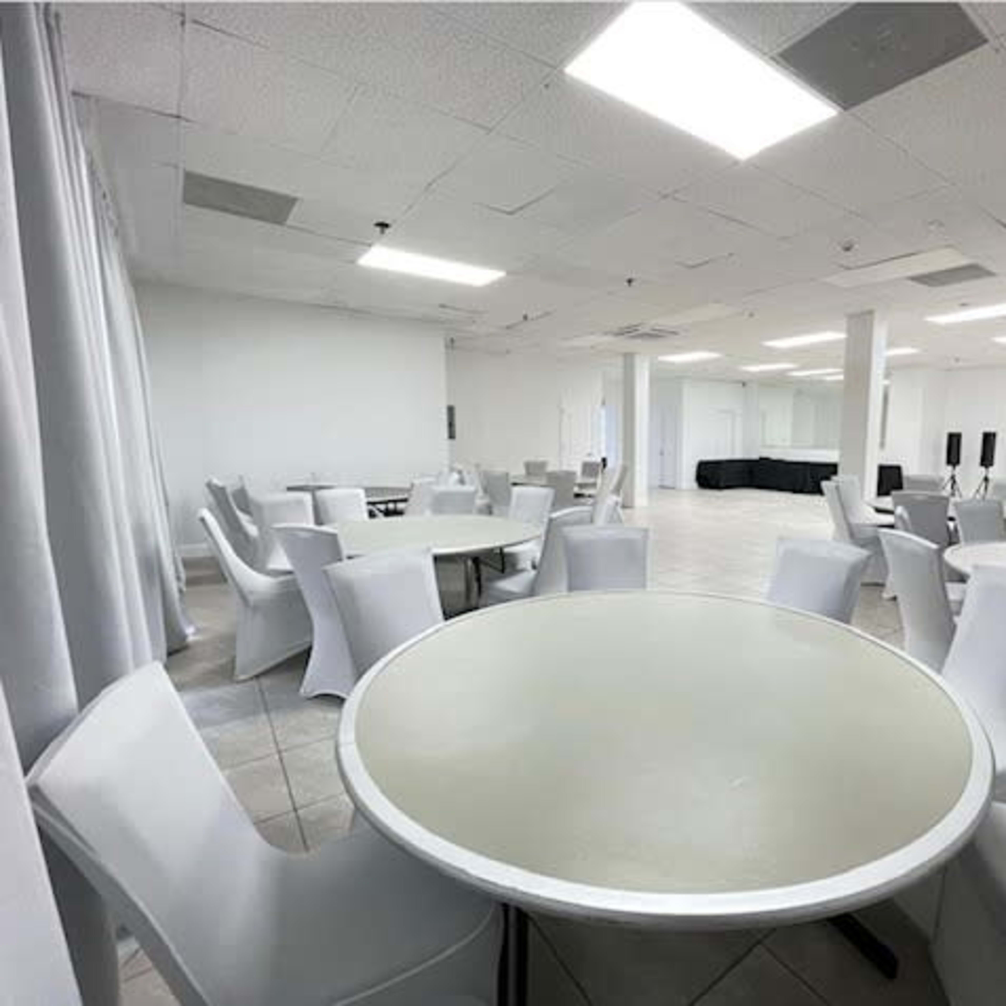 A spacious, empty room with several round tables surrounded by white chairs and bright overhead lights.