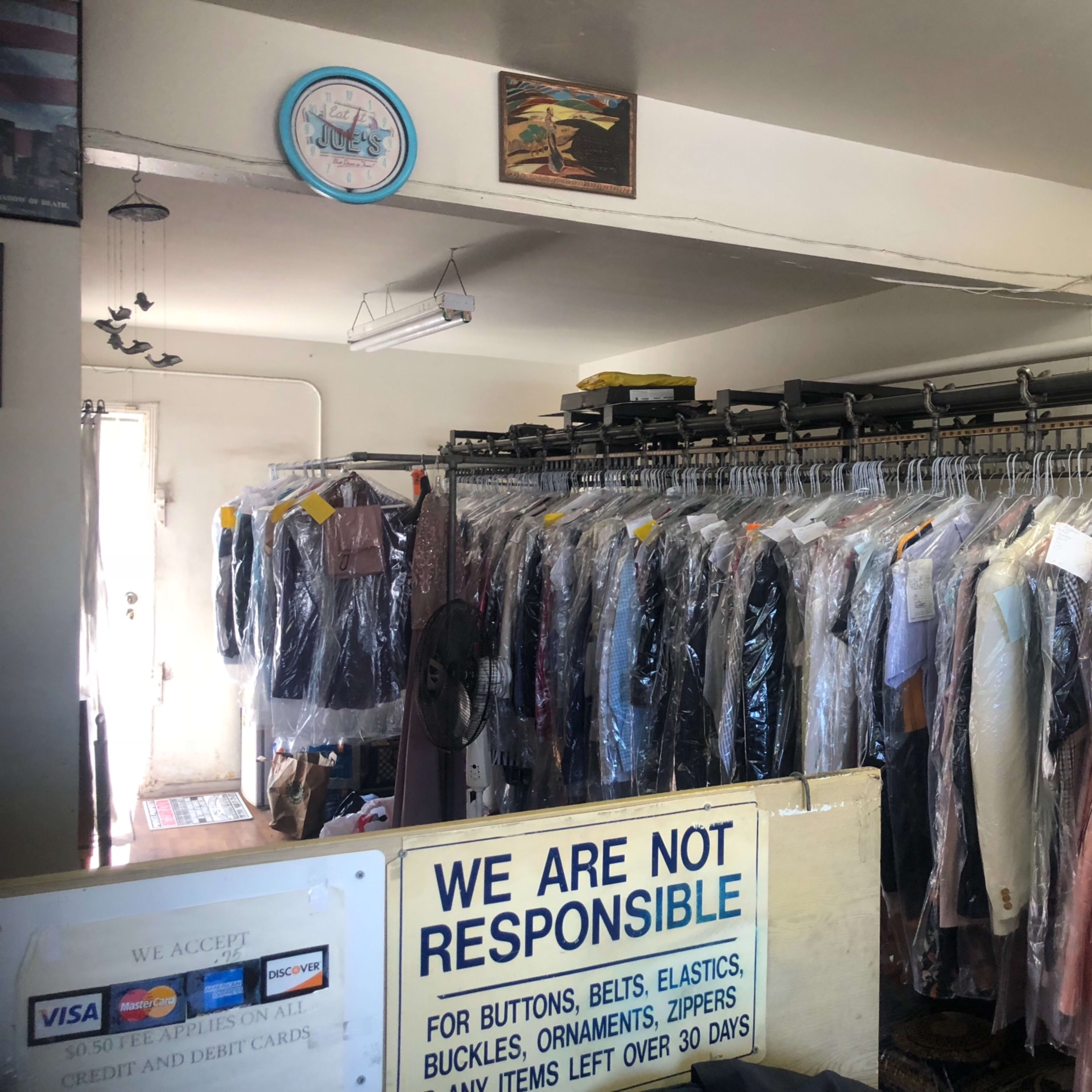 A dry cleaning shop with a rack of hanging garments covered in plastic, and a sign at the counter detailing payment methods and a liability disclaimer.