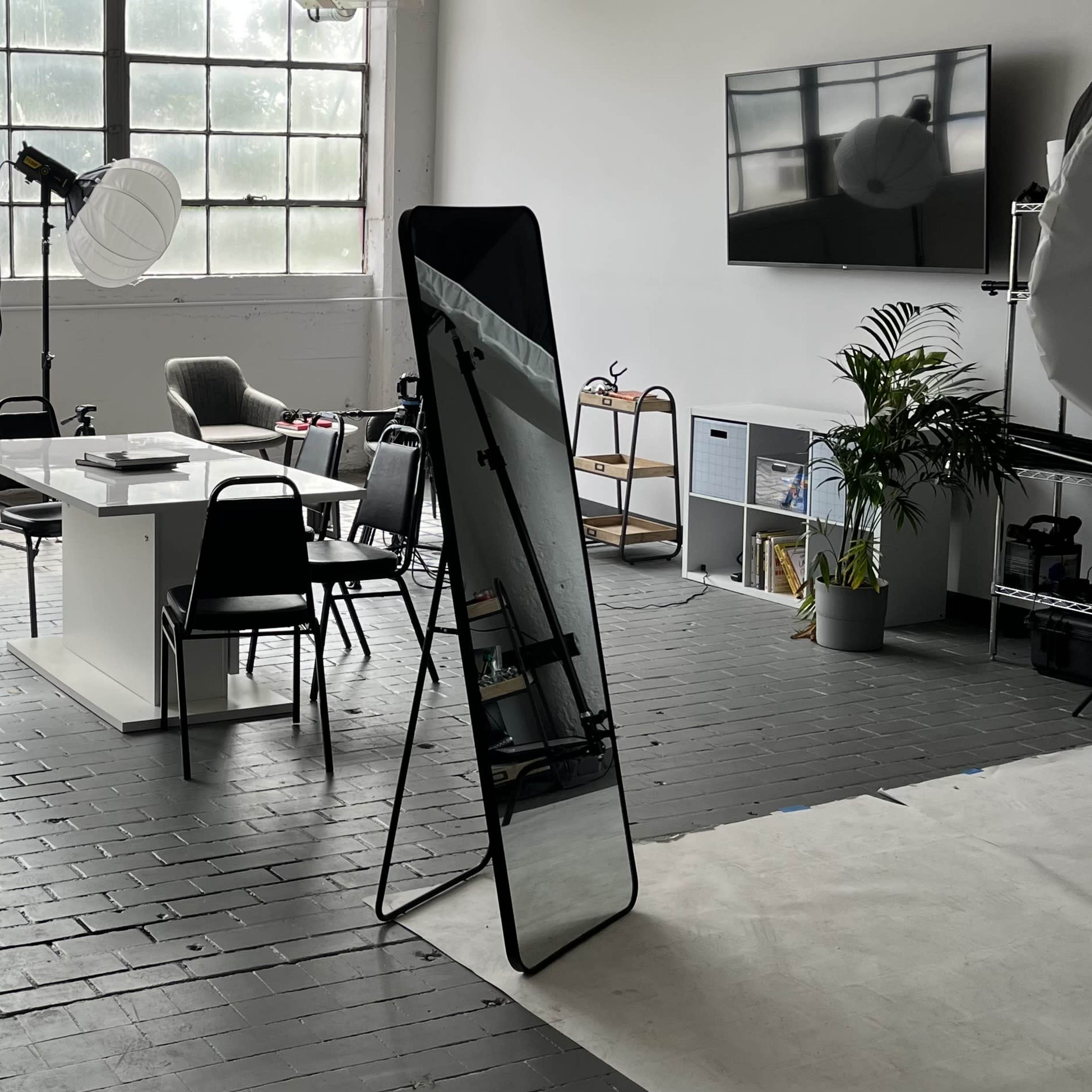A spacious office environment featuring a large window, a white desk with chairs, an oversized mirror, and various plants.