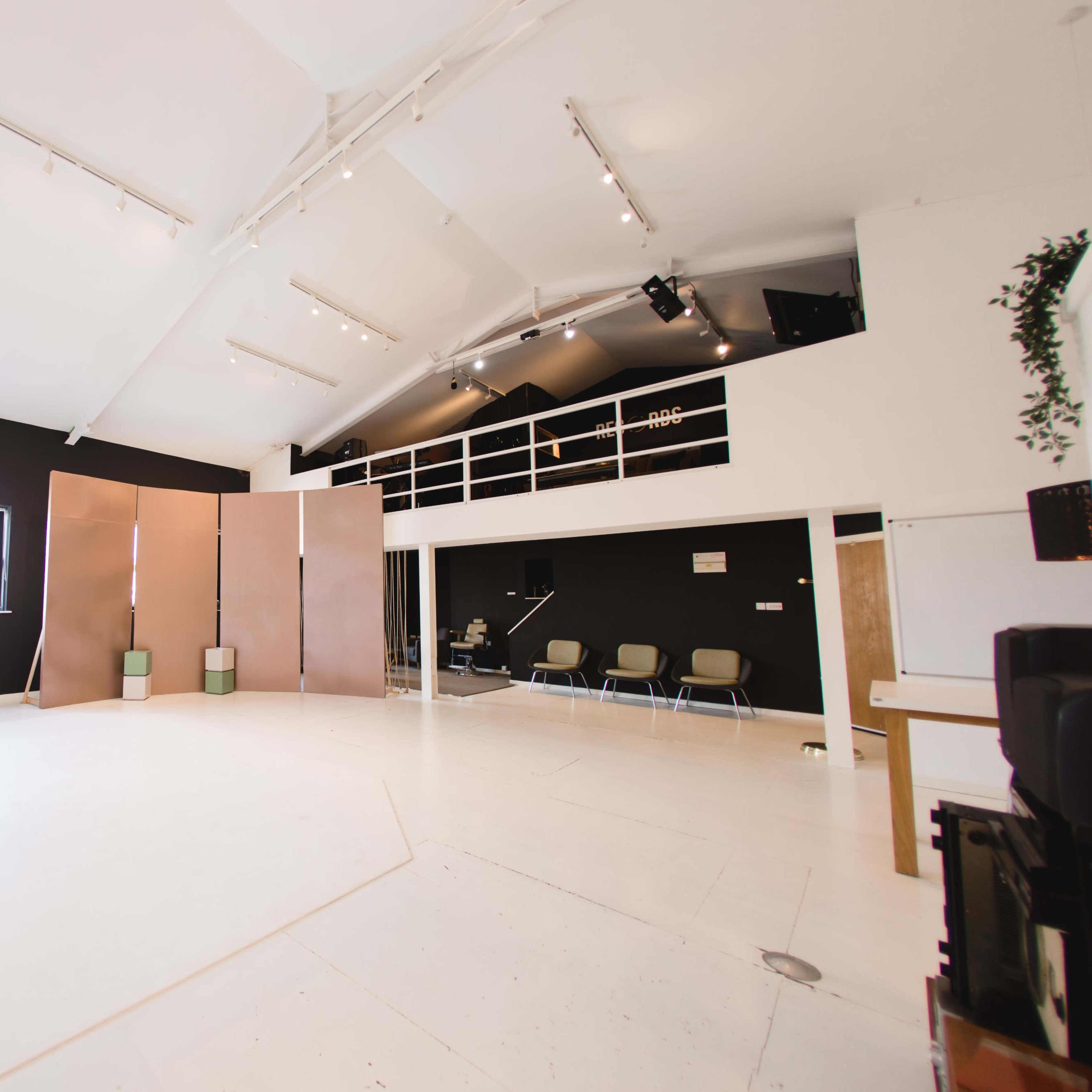 A spacious studio with white floors, black walls, a large window, and a loft area featuring a few chairs and a mannequin in the foreground.