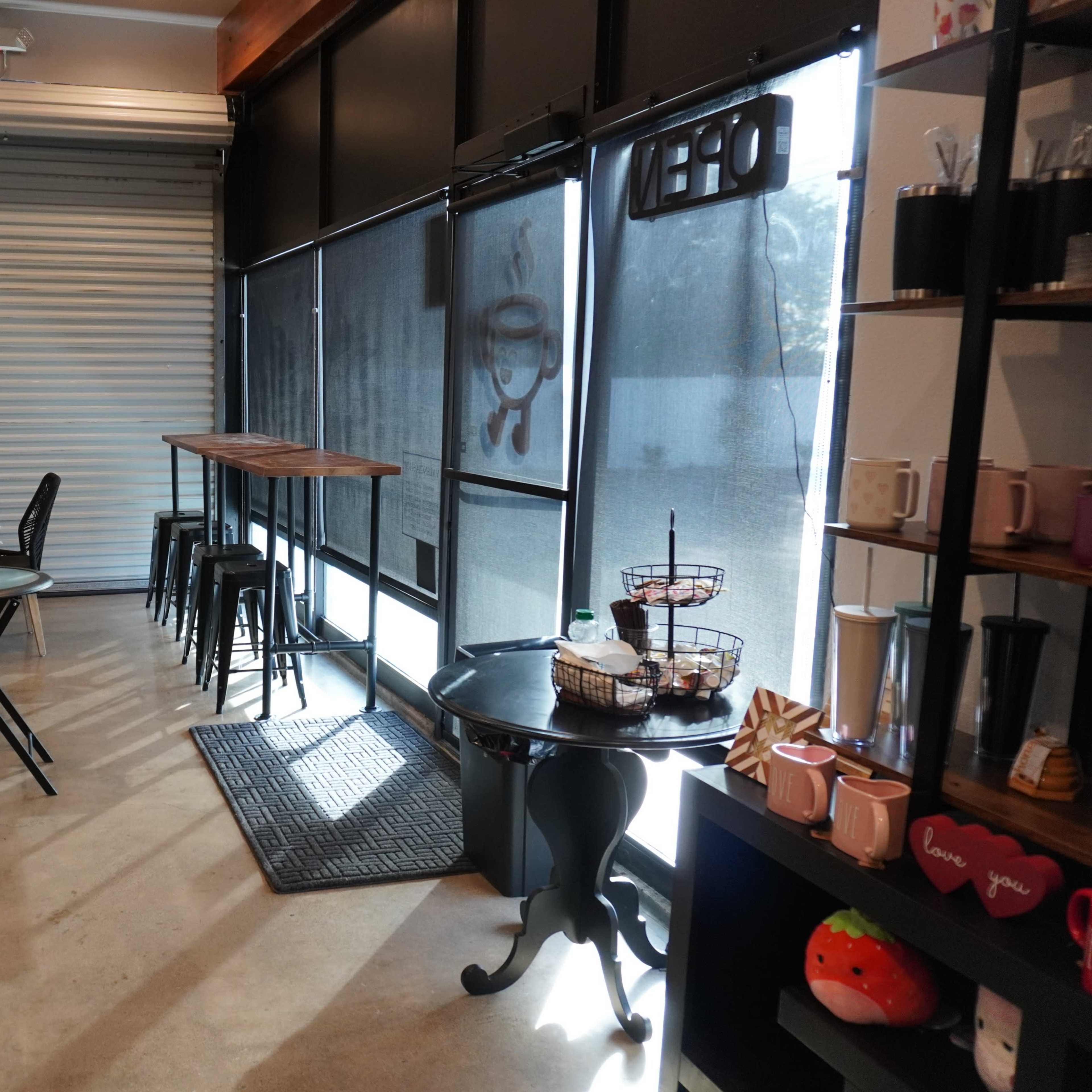 A café interior with black folding chairs and tables, a high table with stools, and a shelf displaying various mugs and kitchen items.