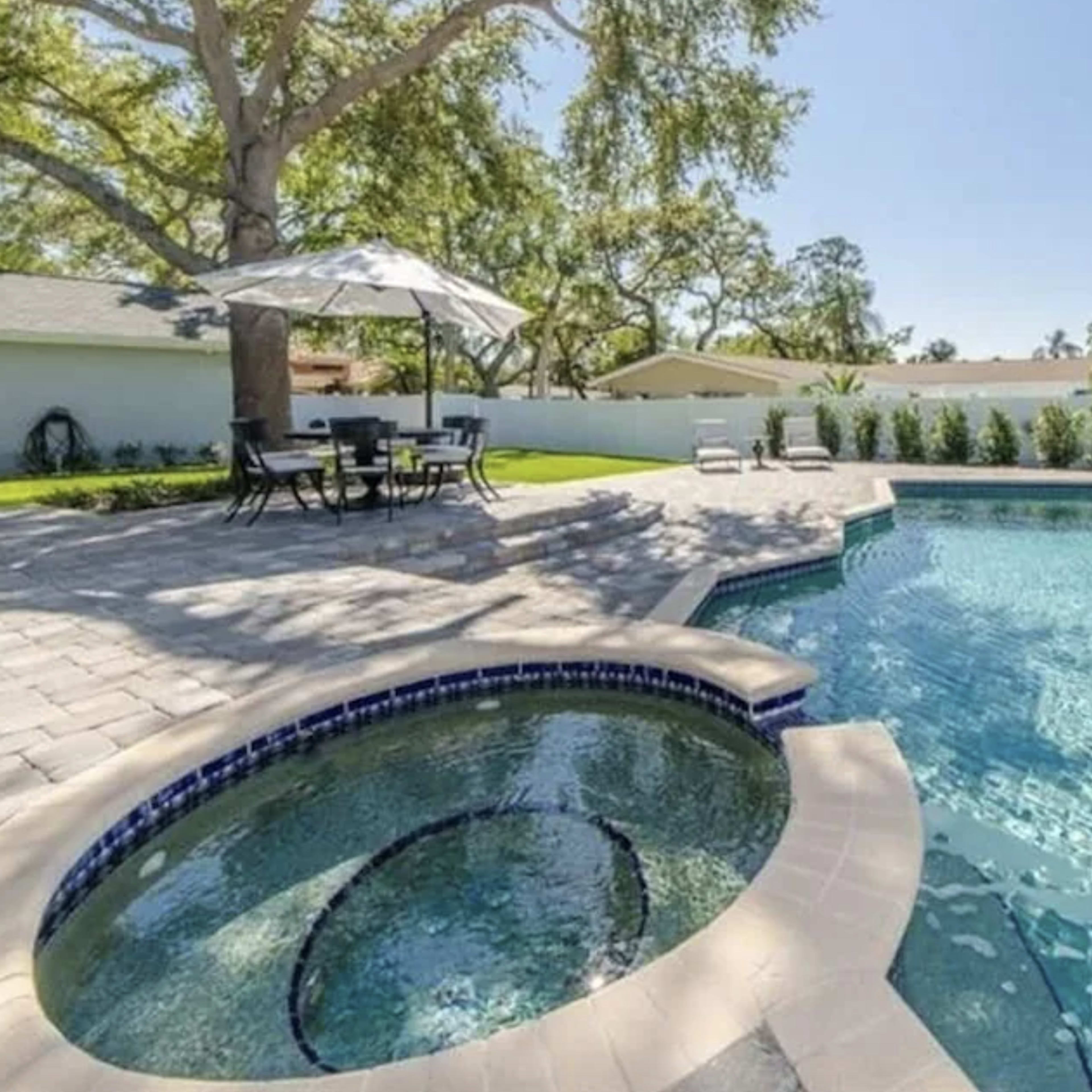 A swimming pool with a hot tub, surrounded by paved stones and lawn furniture under an umbrella in a residential backyard.