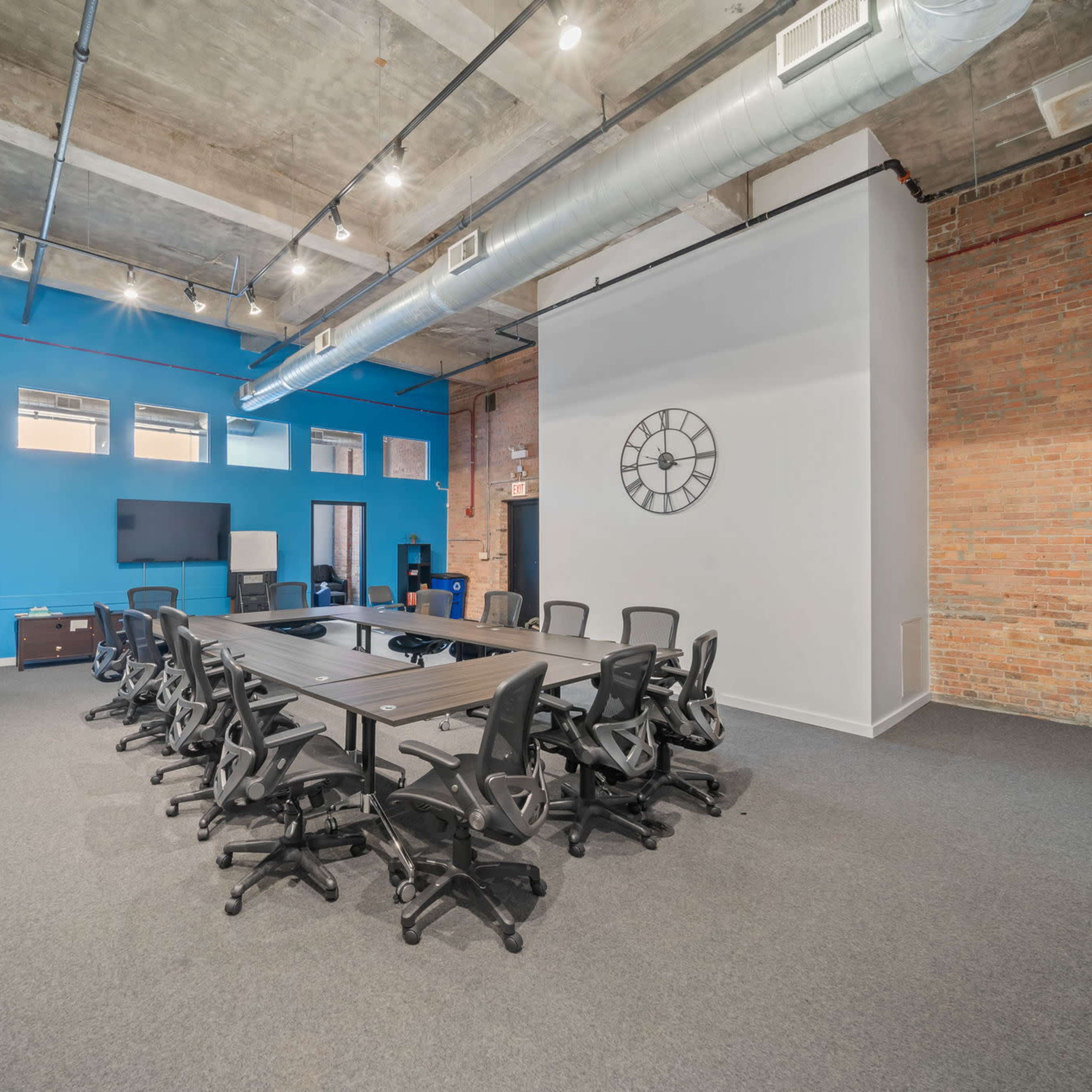 A modern conference room features a large table surrounded by ergonomic chairs, with exposed brick walls and blue accents.