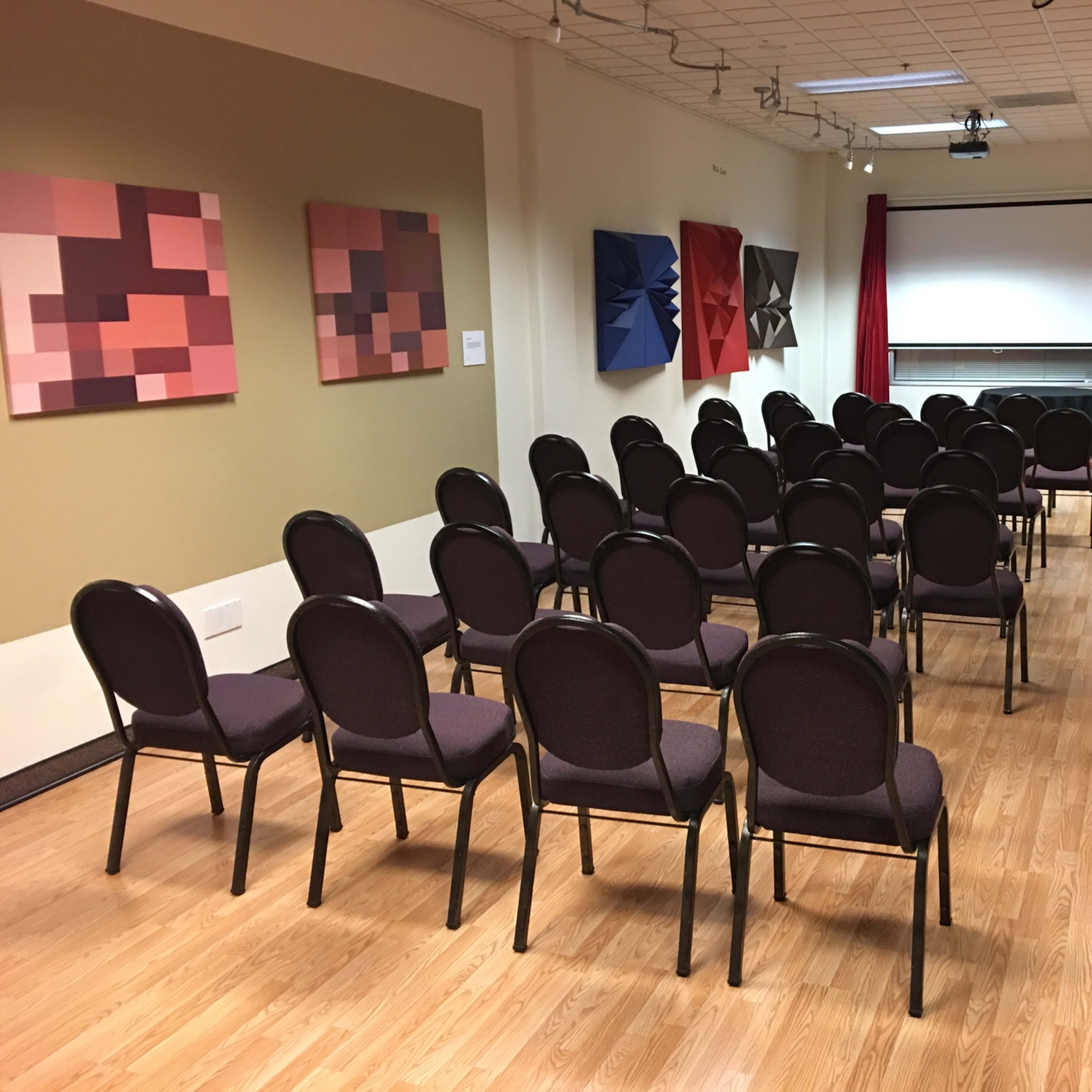 A modestly arranged room featuring rows of chairs facing a screen, with abstract artwork on the walls and red curtains.