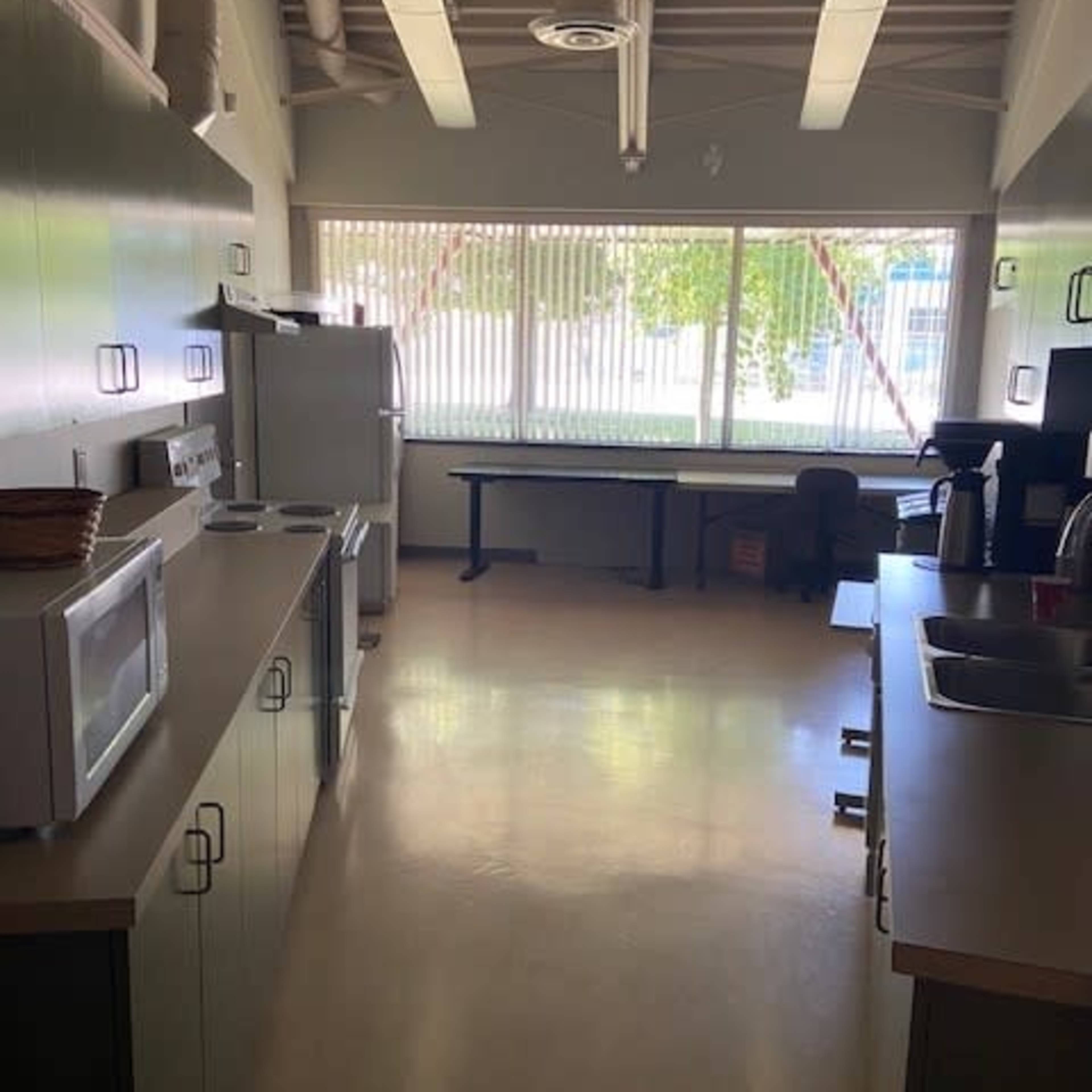 A kitchen area with appliances, including a refrigerator and microwave, adjacent to a workspace with a table and chairs, and large windows allowing natural light.