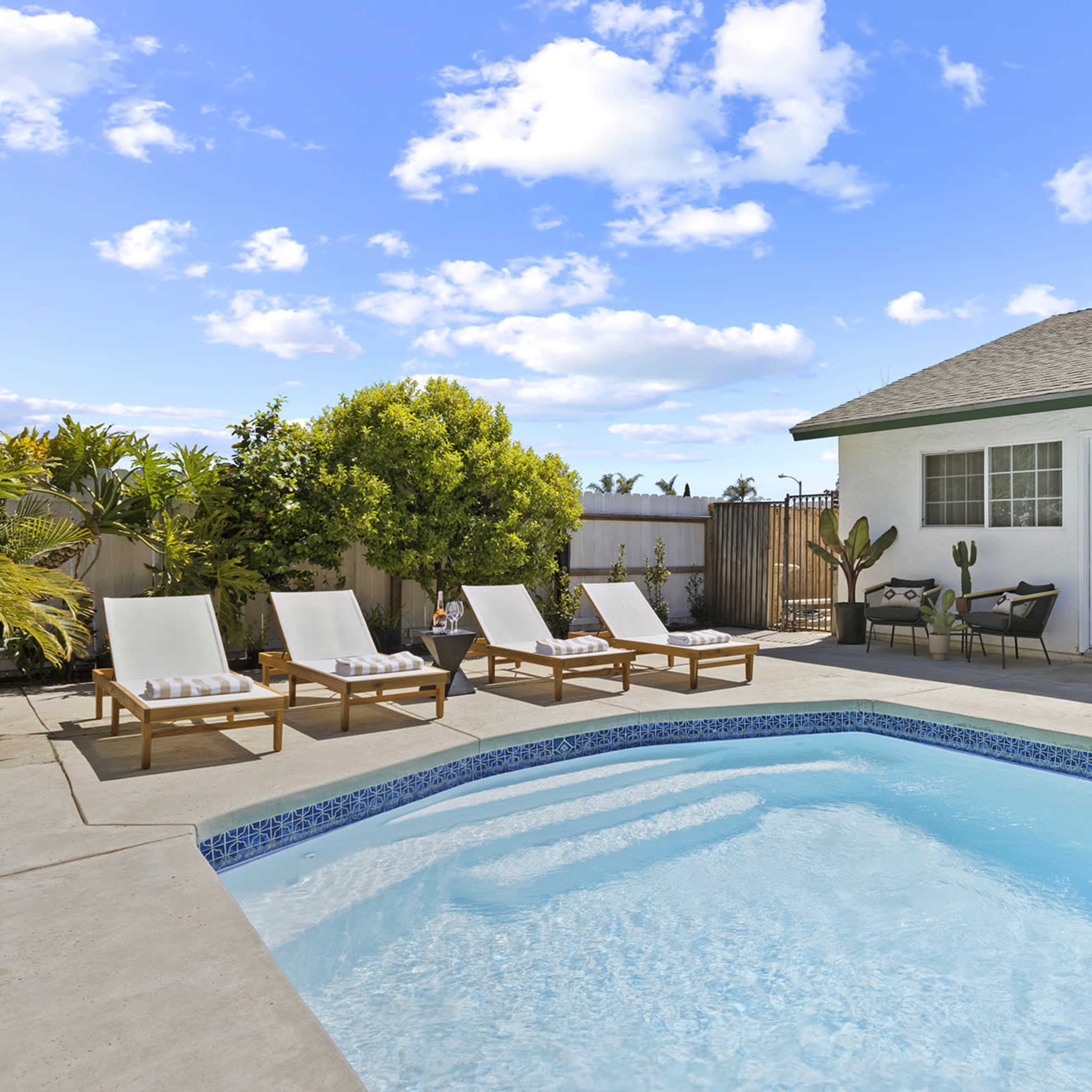 A swimming pool surrounded by lounge chairs and potted plants, with a house in the background under a partly cloudy sky.