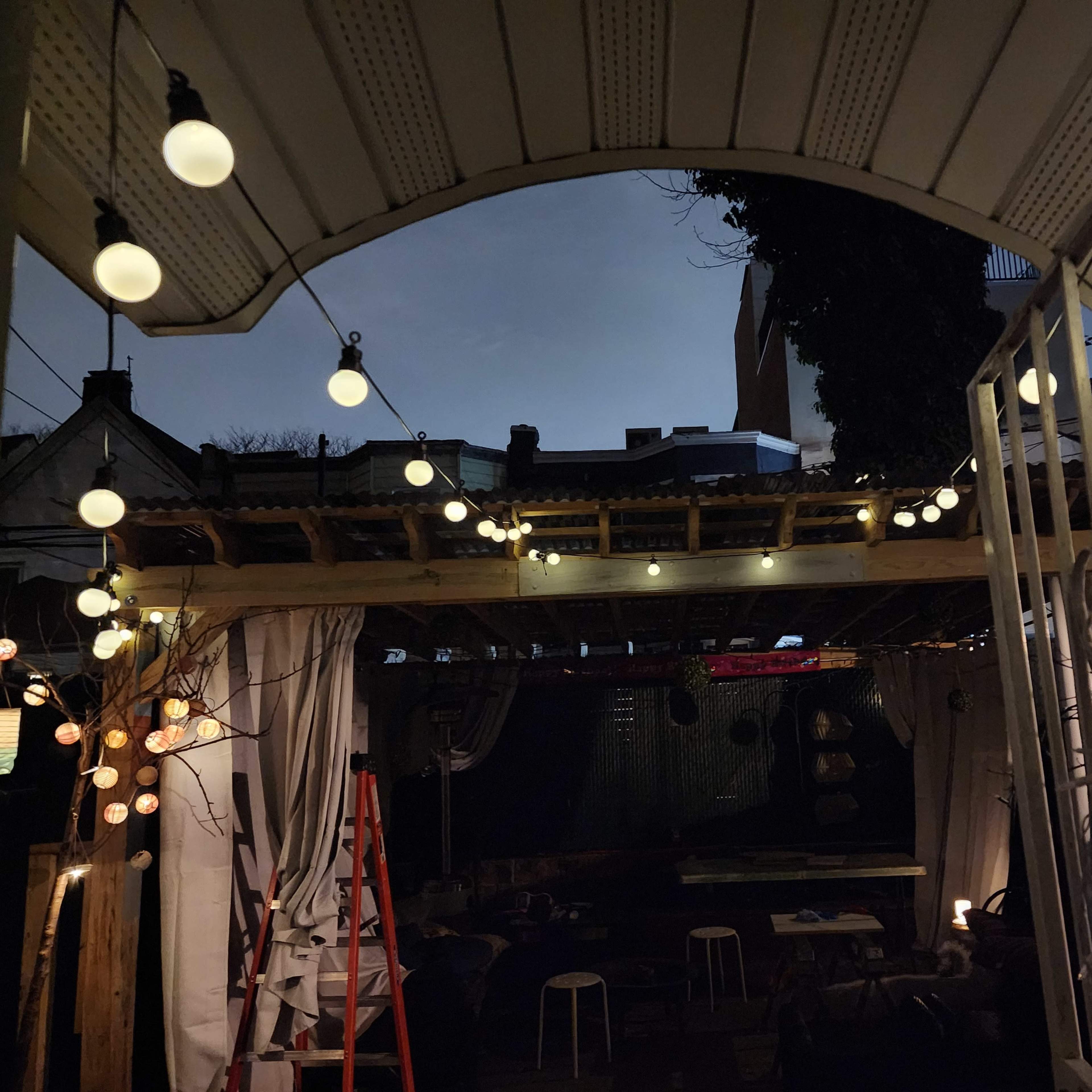 An outdoor patio area illuminated by hanging lights under a cloudy night sky, with a ladder positioned nearby.