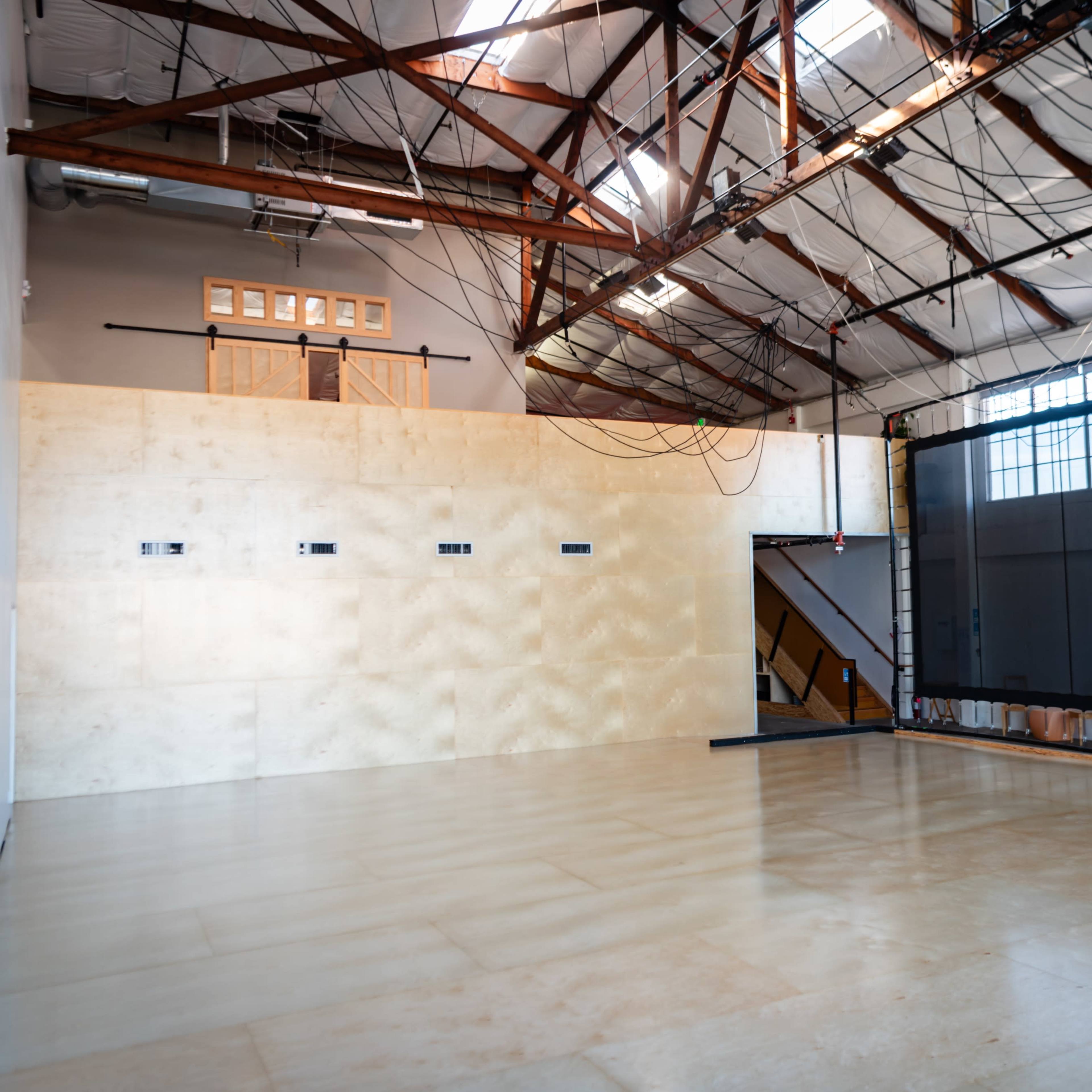 An industrial-style interior with a high ceiling, wooden beams, and a large empty wall opposite a dark projection screen.