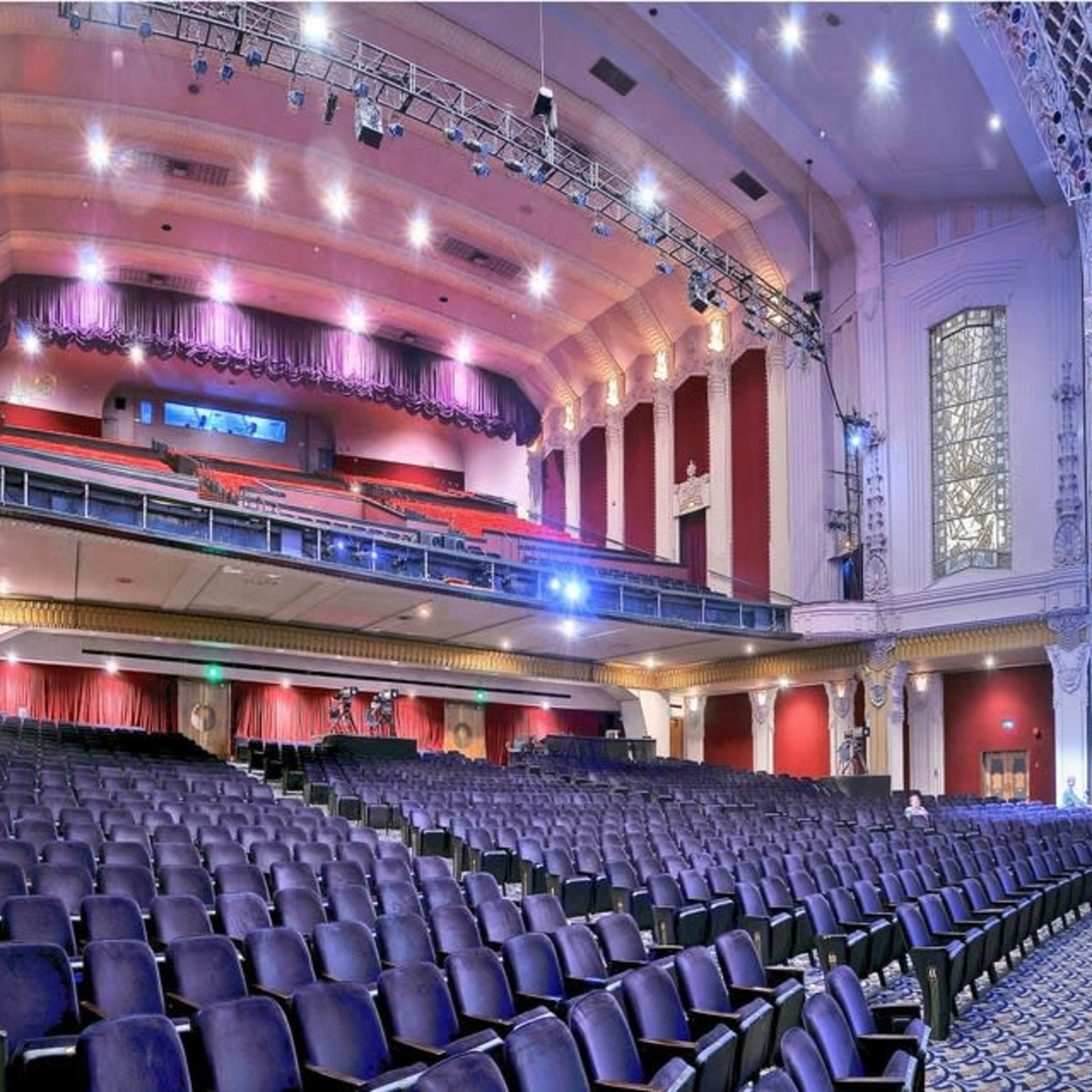 A spacious theater interior with a central stage, rows of empty seats, and ornate architectural details on the walls and ceiling.