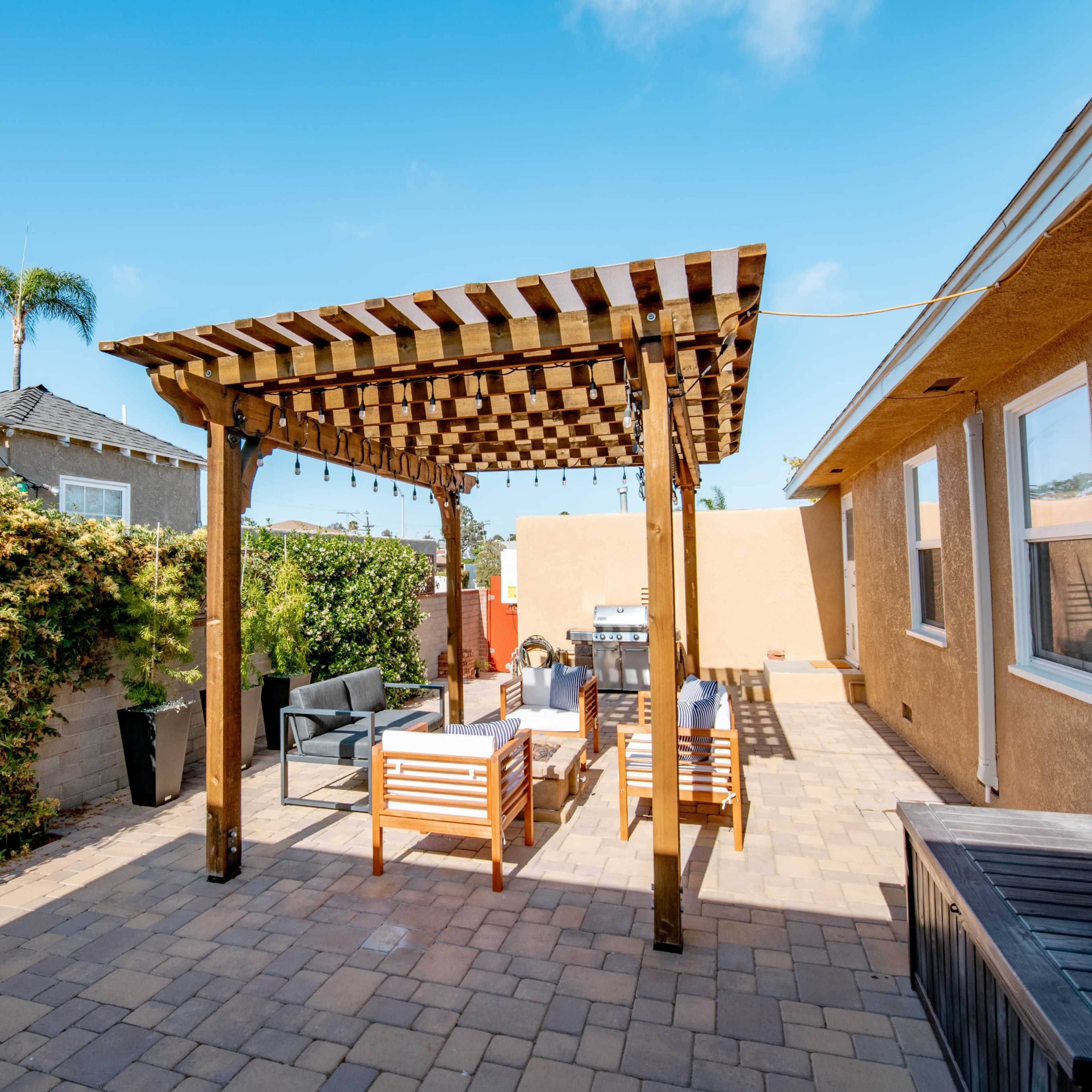 A patio area with a wooden pergola, seating arrangements, and paver stones surrounded by greenery.