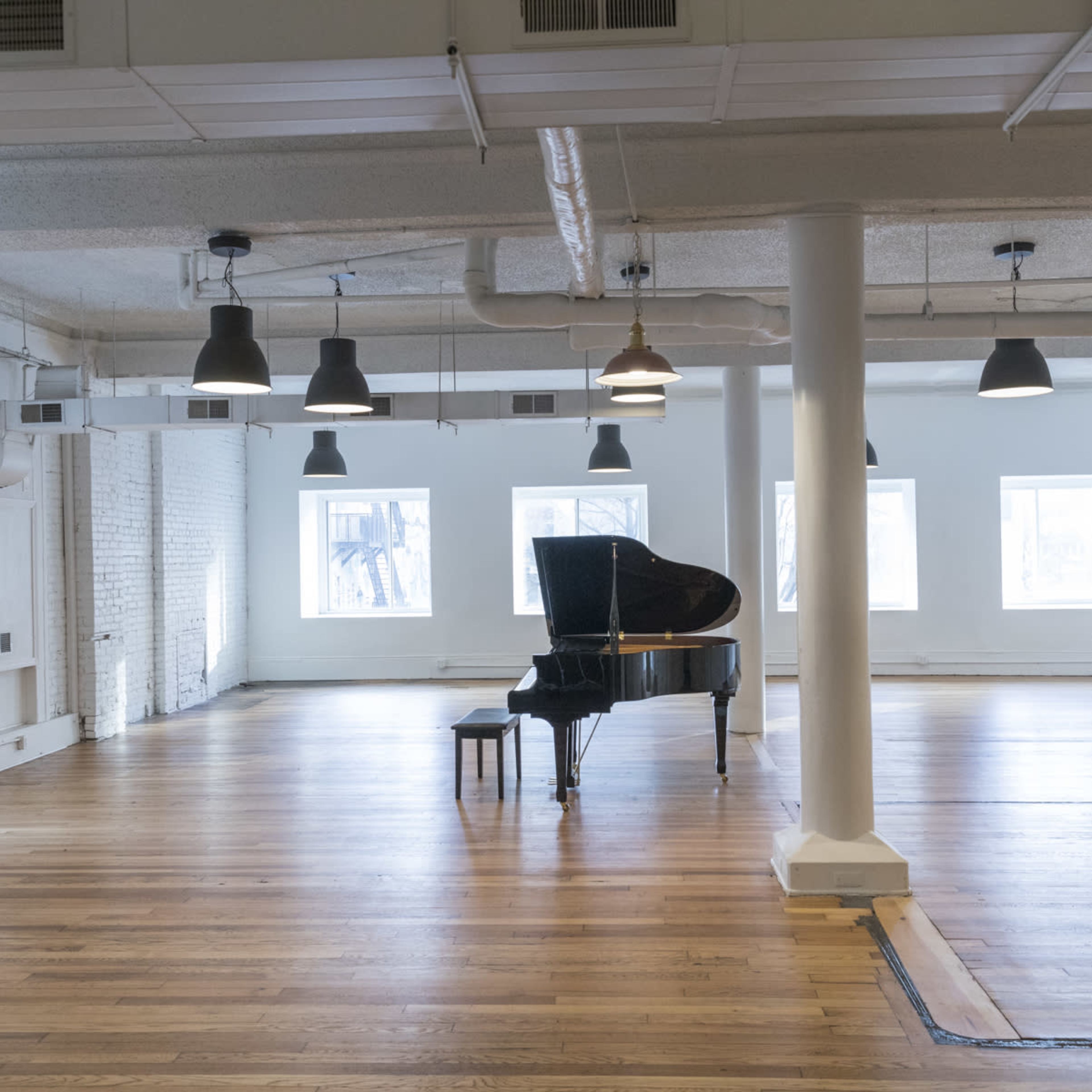 A grand piano sits alone in the center of a spacious, empty room with wooden floors and multiple windows.