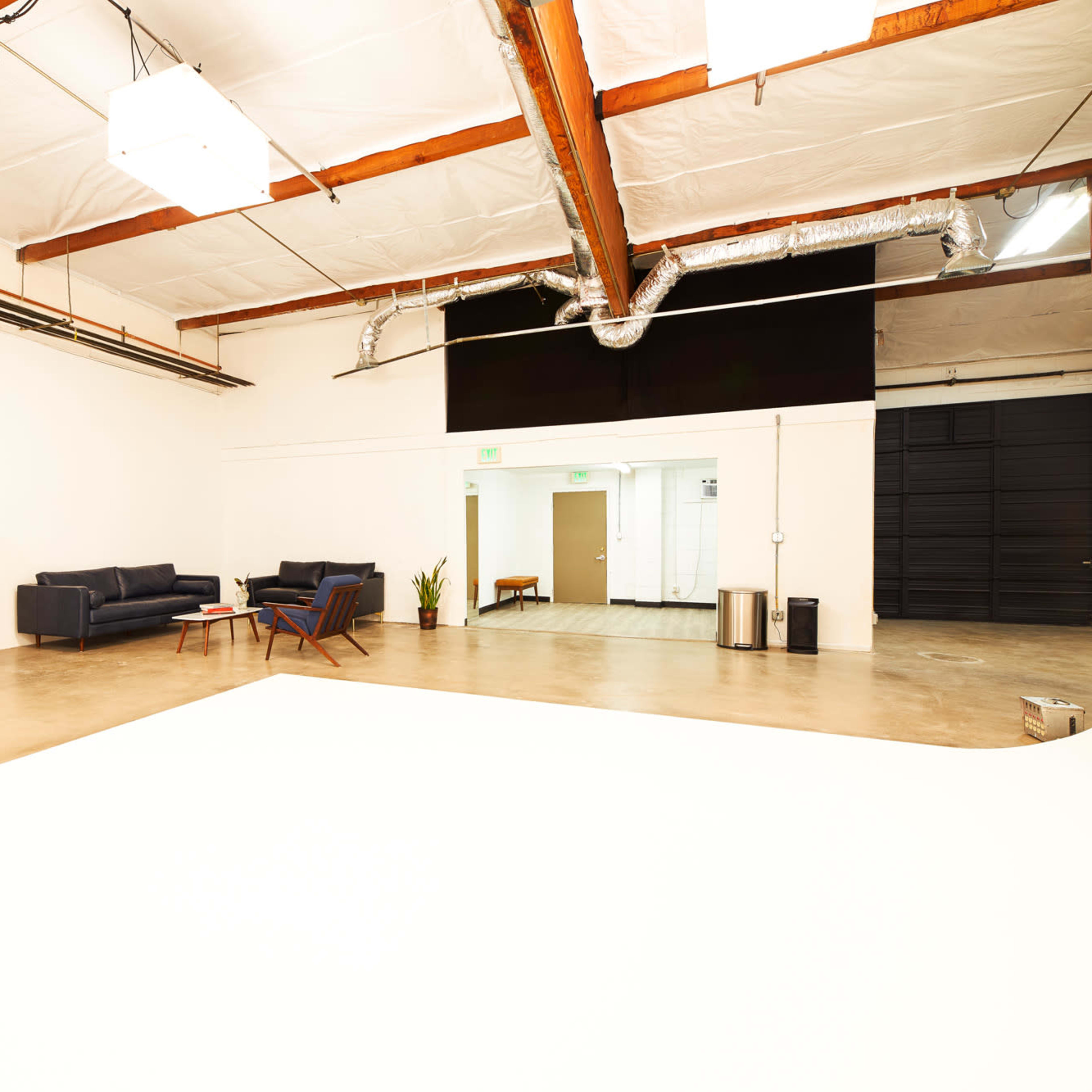 A spacious, well-lit studio features a sectional sofa, a seating area, a kitchenette with a microwave, and adjacent doors leading to another room.