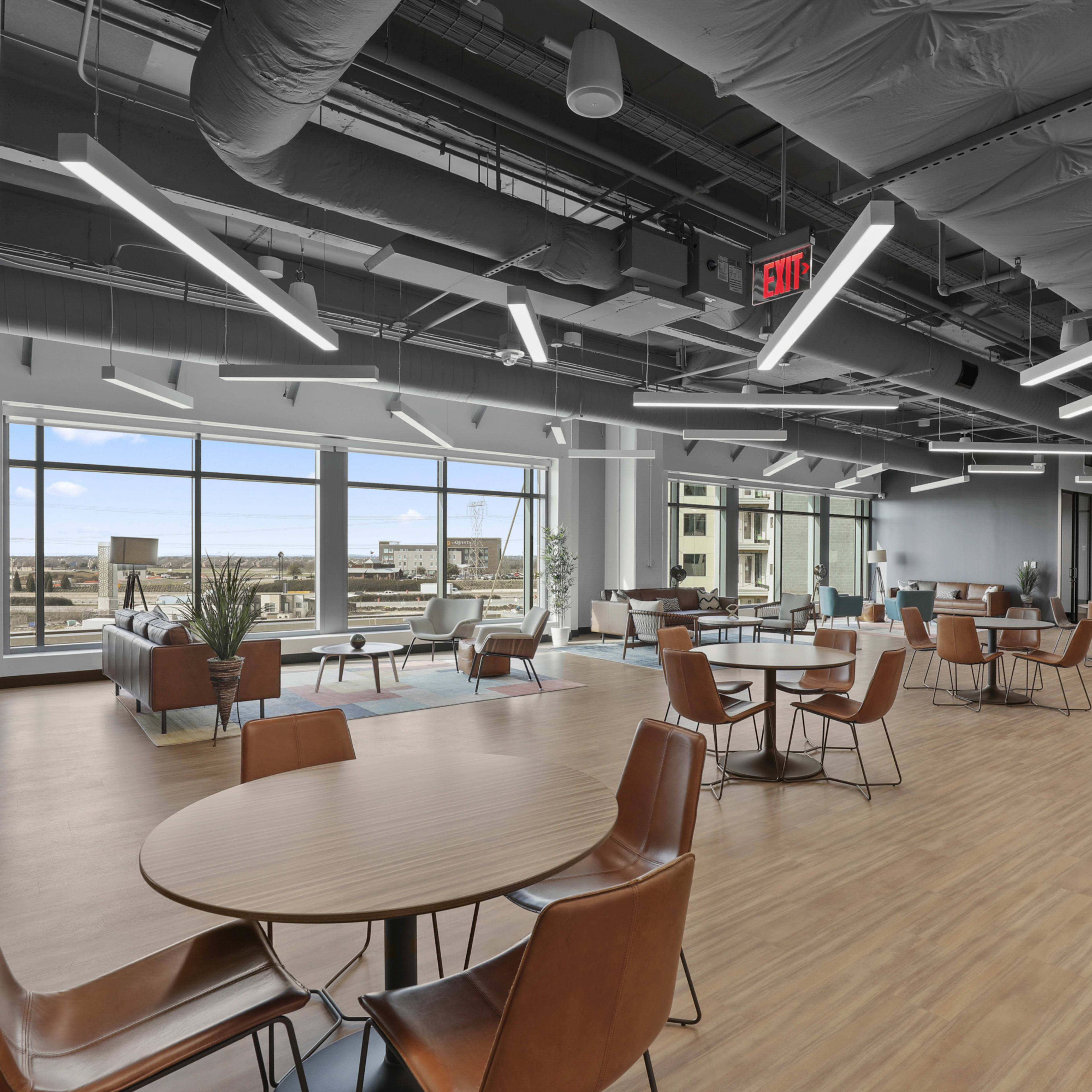 A modern office lounge area with wooden tables and chairs, large windows, and a spacious layout.
