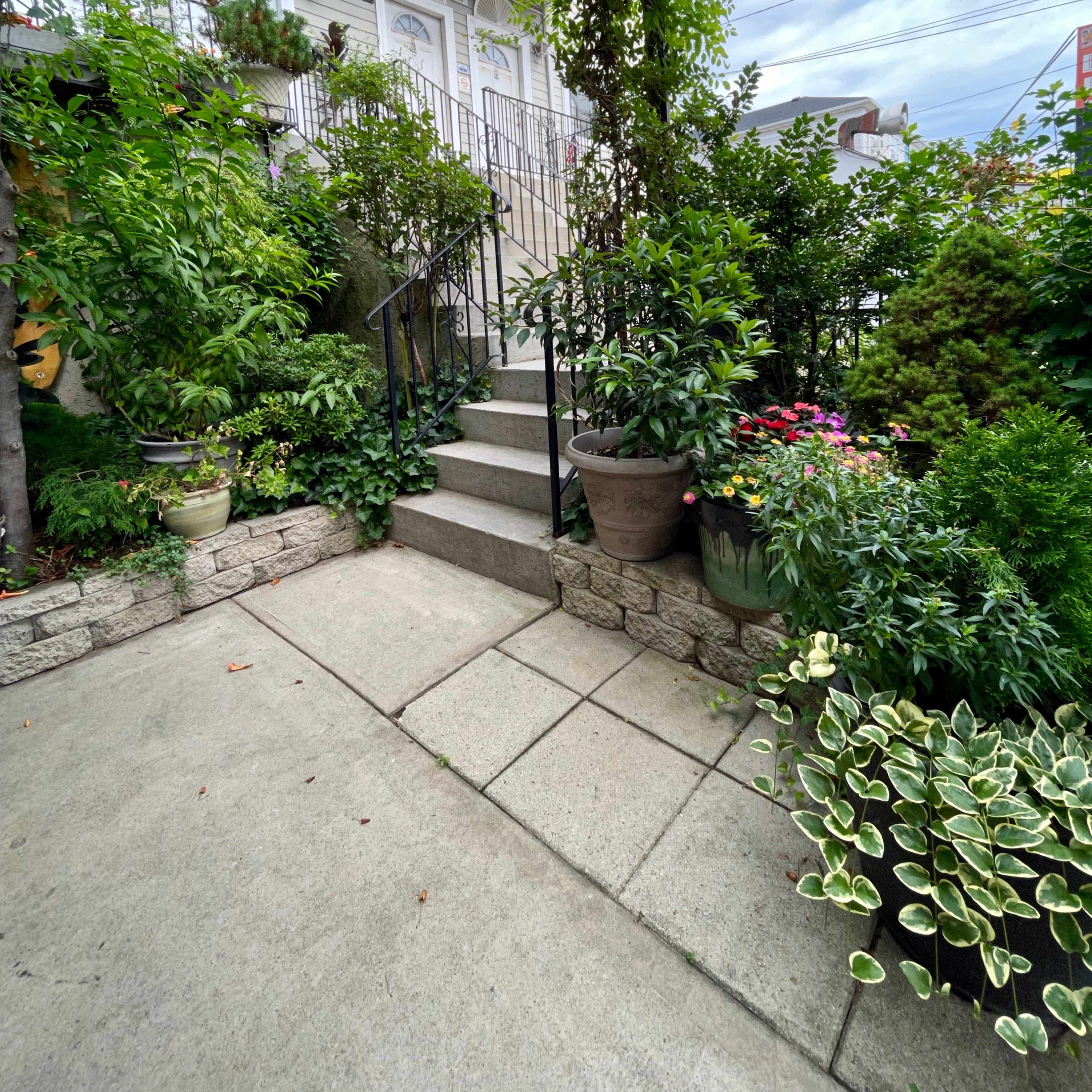 A stone pathway leads to a set of stairs flanked by various potted plants and greenery.
