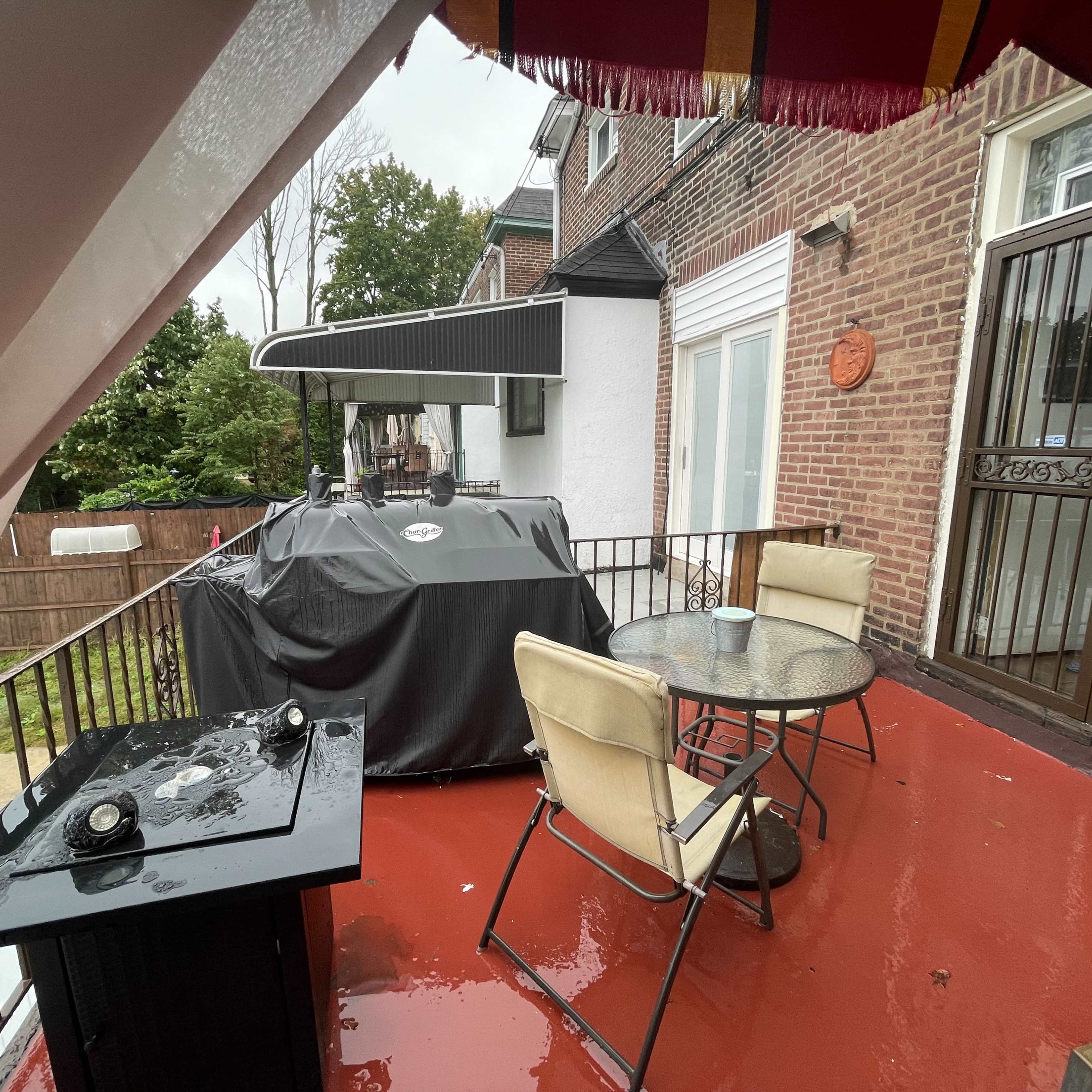 A covered patio features a table with two chairs and a barbecue grill, set against a backdrop of wet surfaces on a cloudy day.