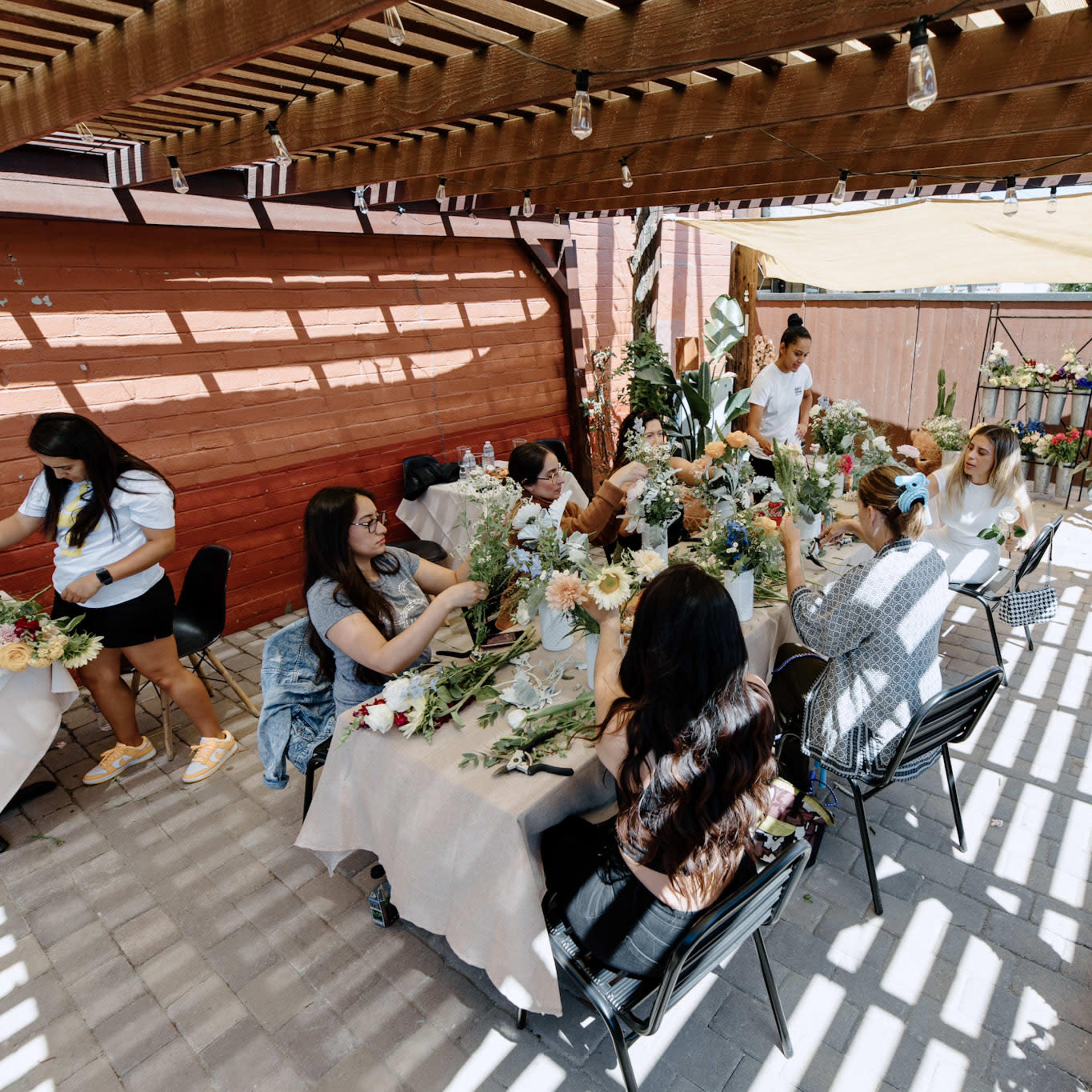A group of people is gathered around tables in a shaded outdoor space, arranging flowers for a craft activity.