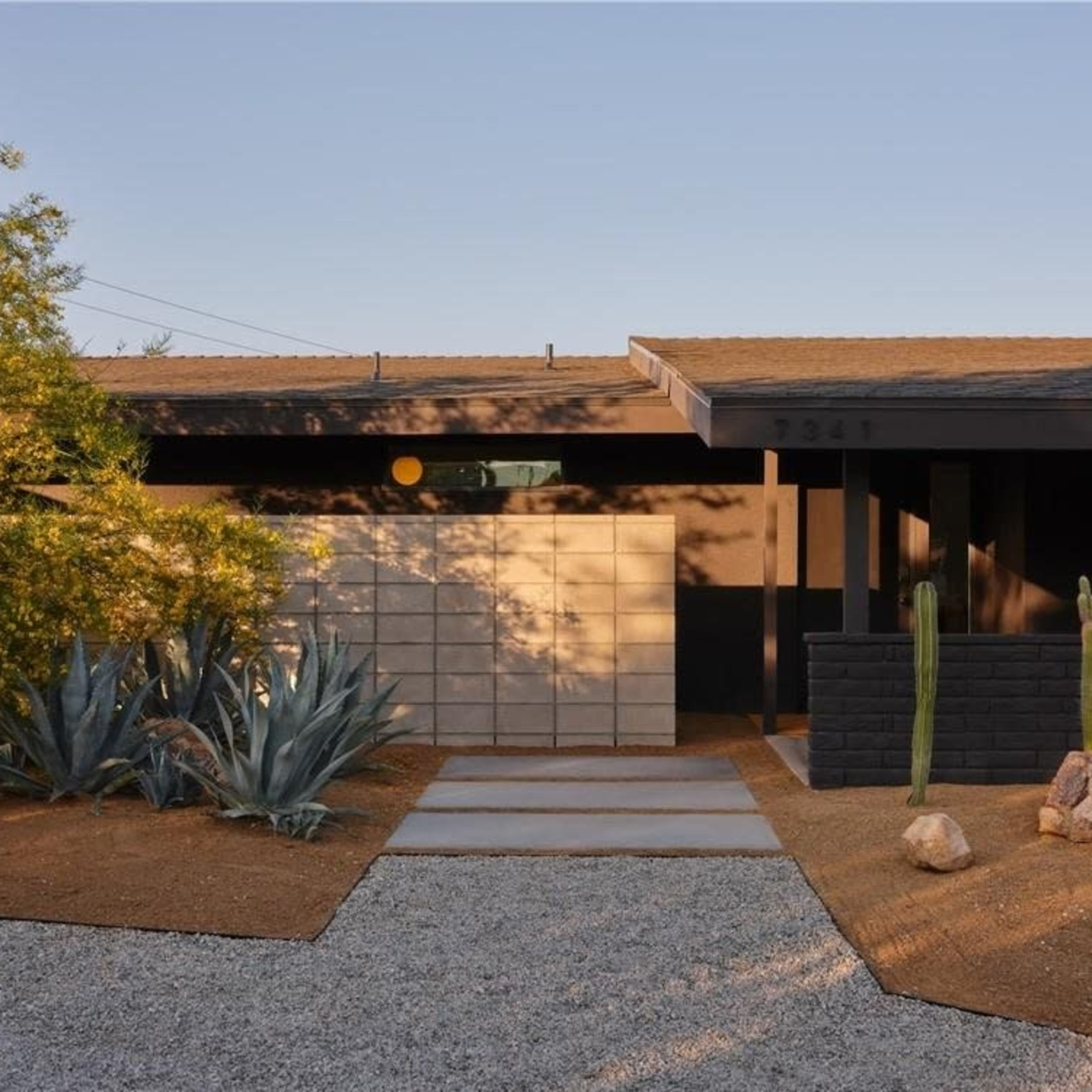 A modern house with a textured black facade, a stone walkway leading to the entrance, and desert landscaping featuring cacti and agave plants.