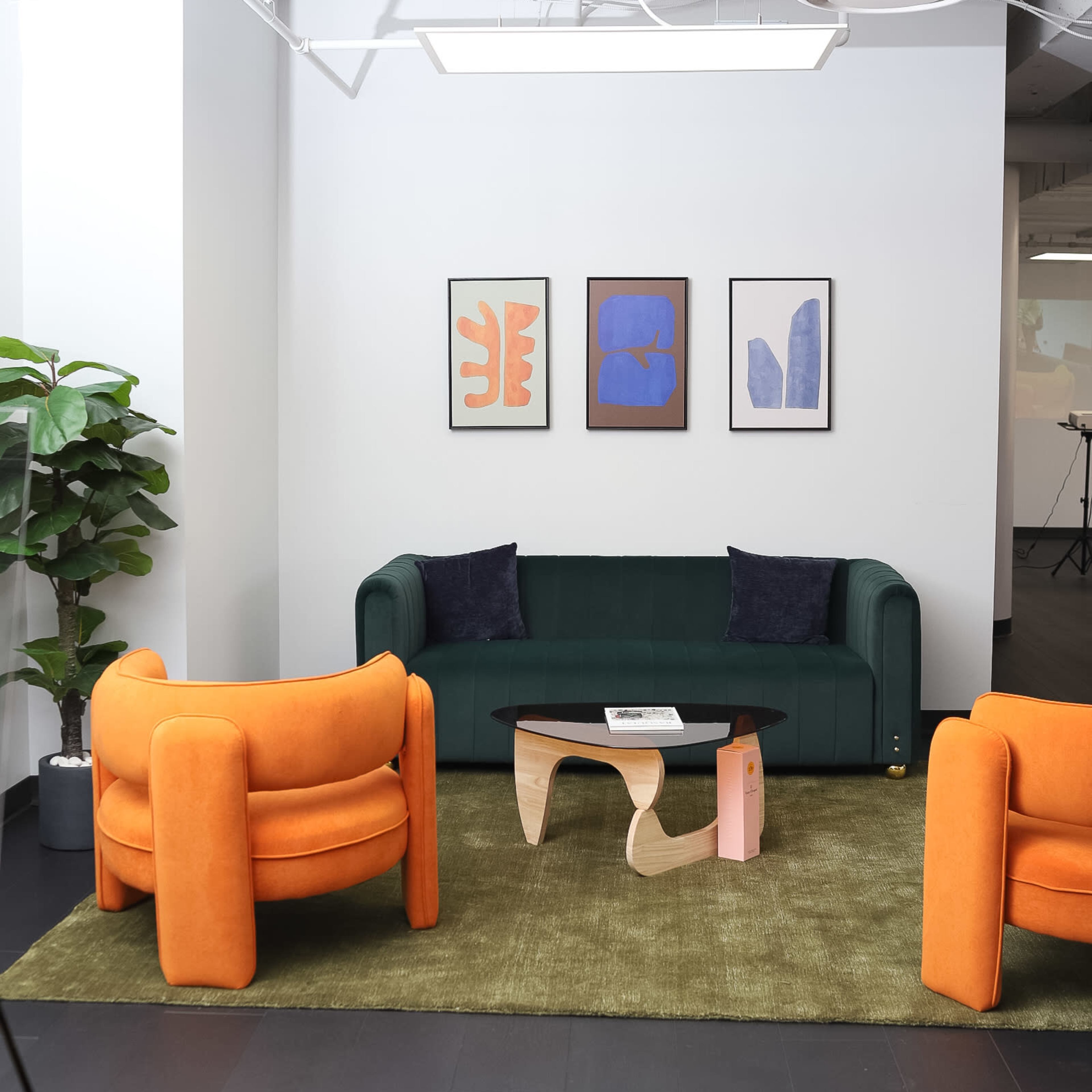 A modern interior with a green sofa, two orange armchairs, a wooden coffee table, and three framed artworks on the wall.