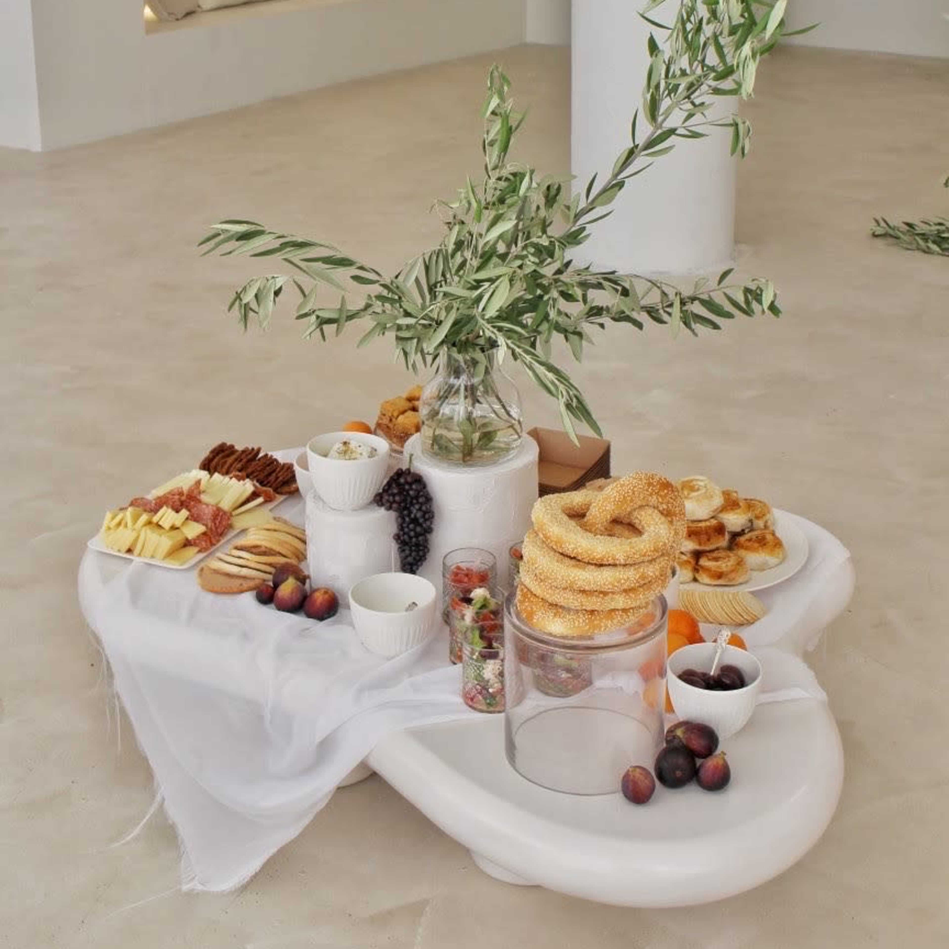 A minimalist food display features an assortment of cheeses, fruits, and pastries arranged on a white table with a decorative olive branch in a spacious, light-filled area.