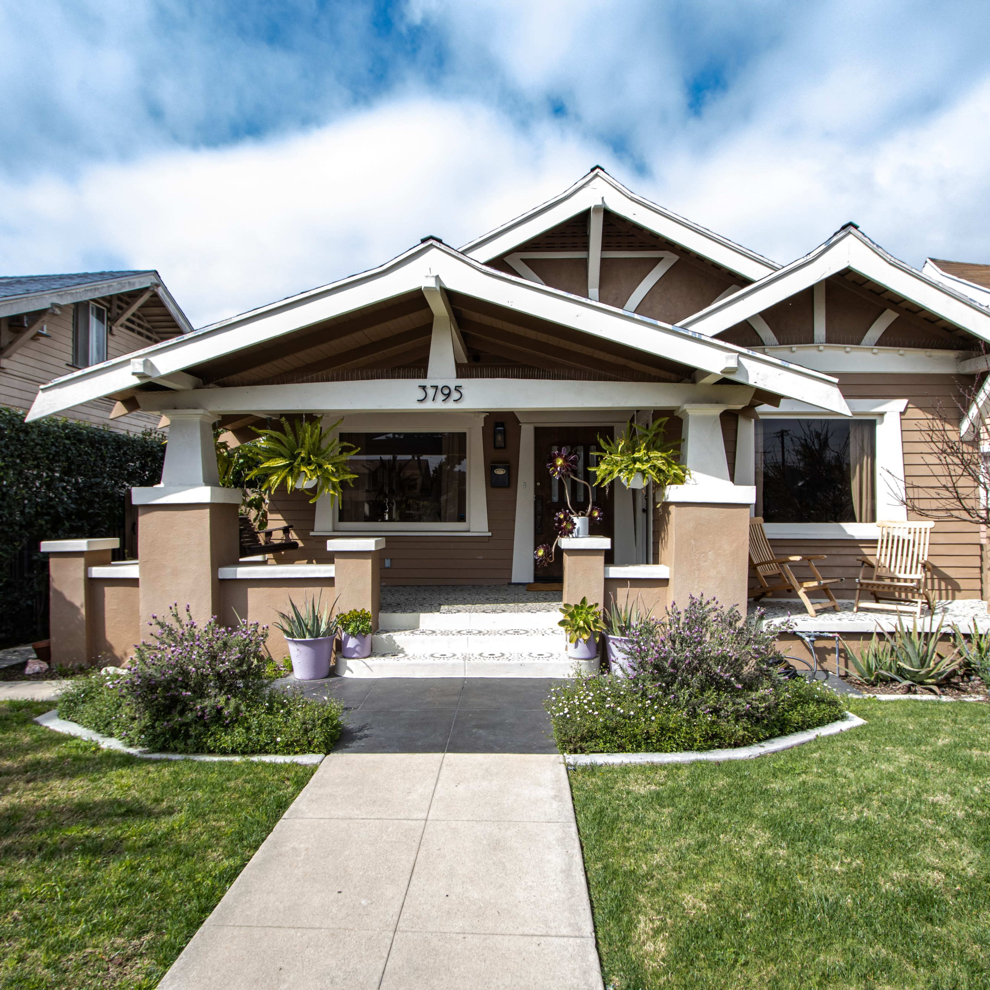 A bungalow-style house with a front porch, surrounded by landscaped greenery and a clear pathway leading to the entrance.