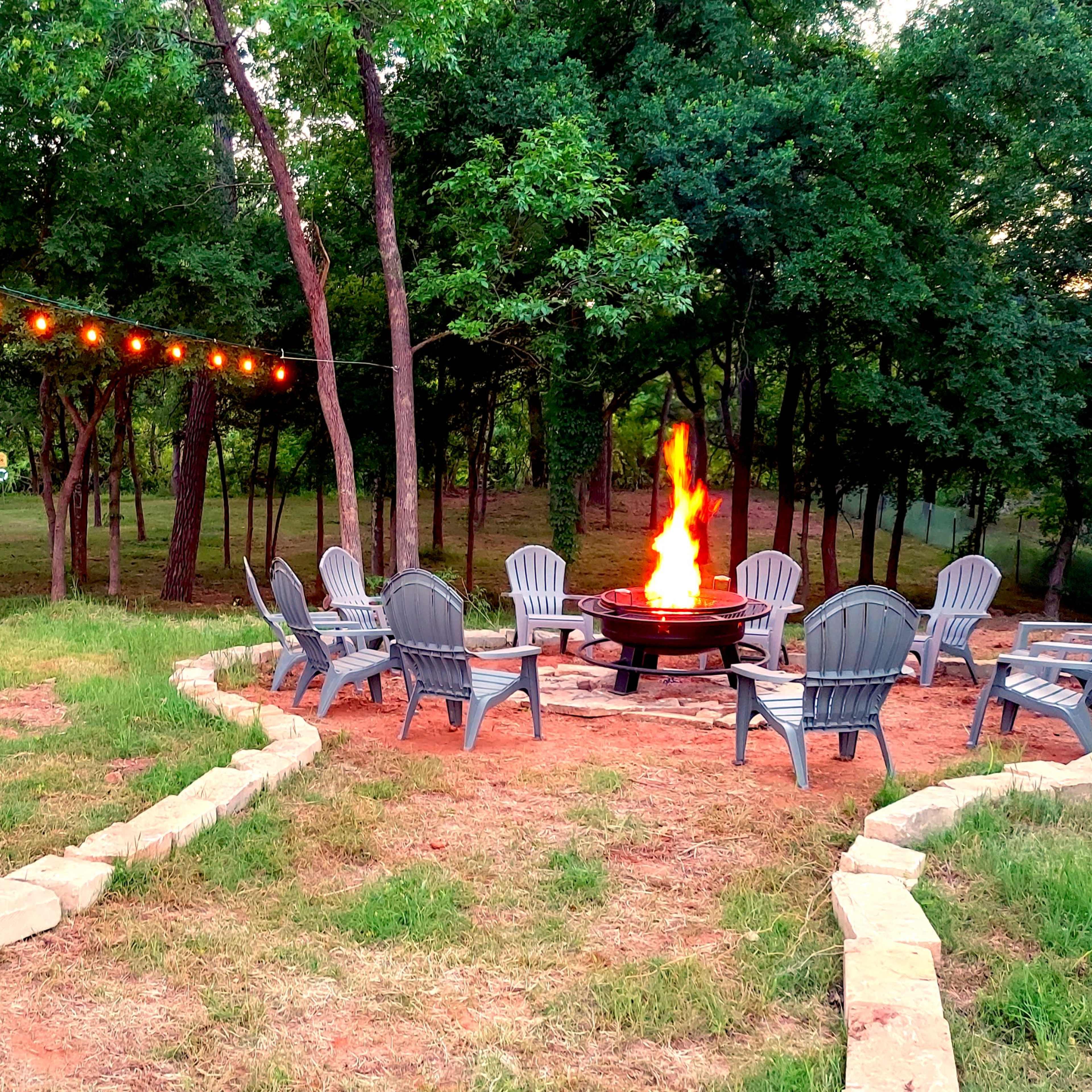 A fire pit surrounded by adirondack chairs is illuminated by string lights in a wooded outdoor setting.