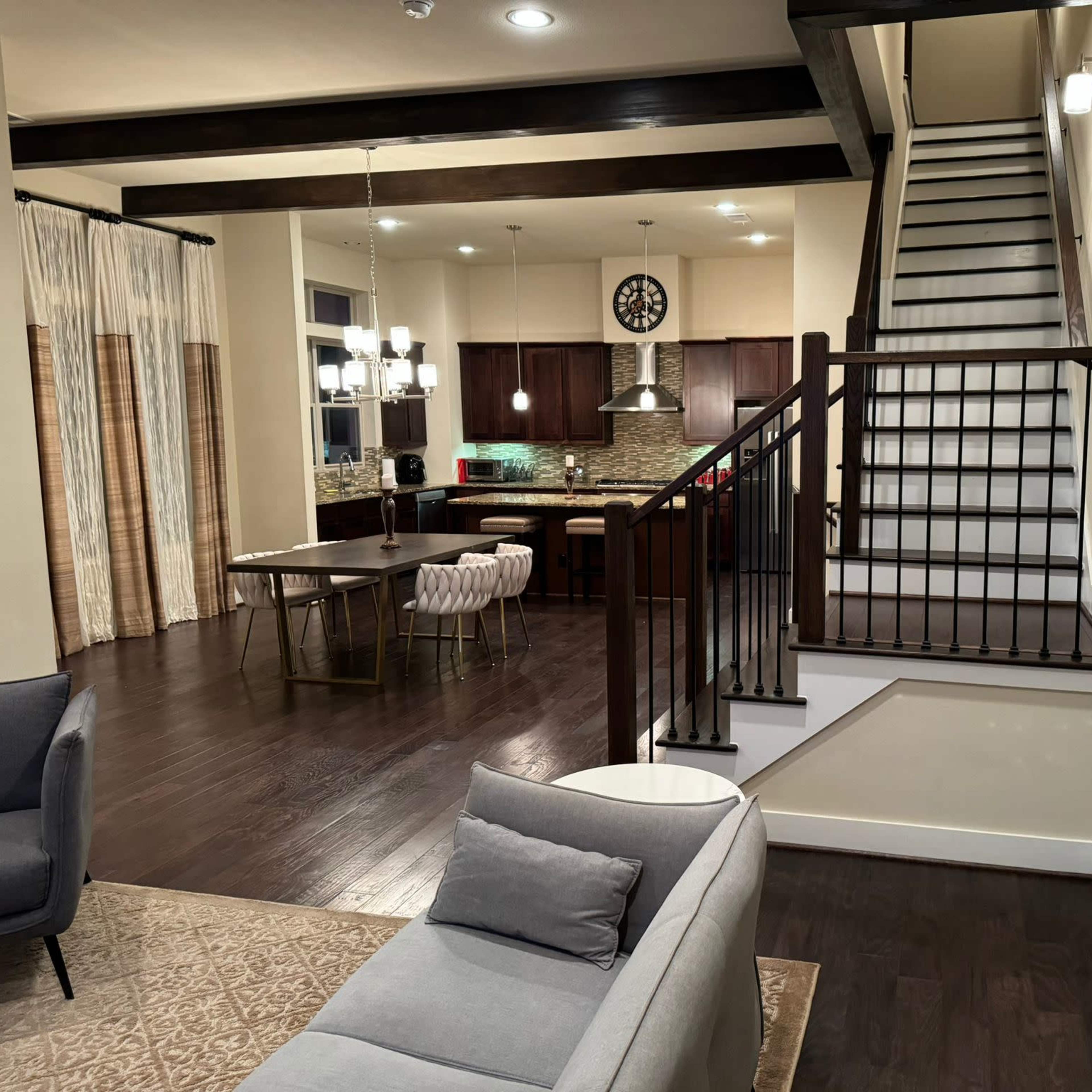 A modern living space with a staircase on the right, a dining area with a round table in the center, and a kitchen in the background.