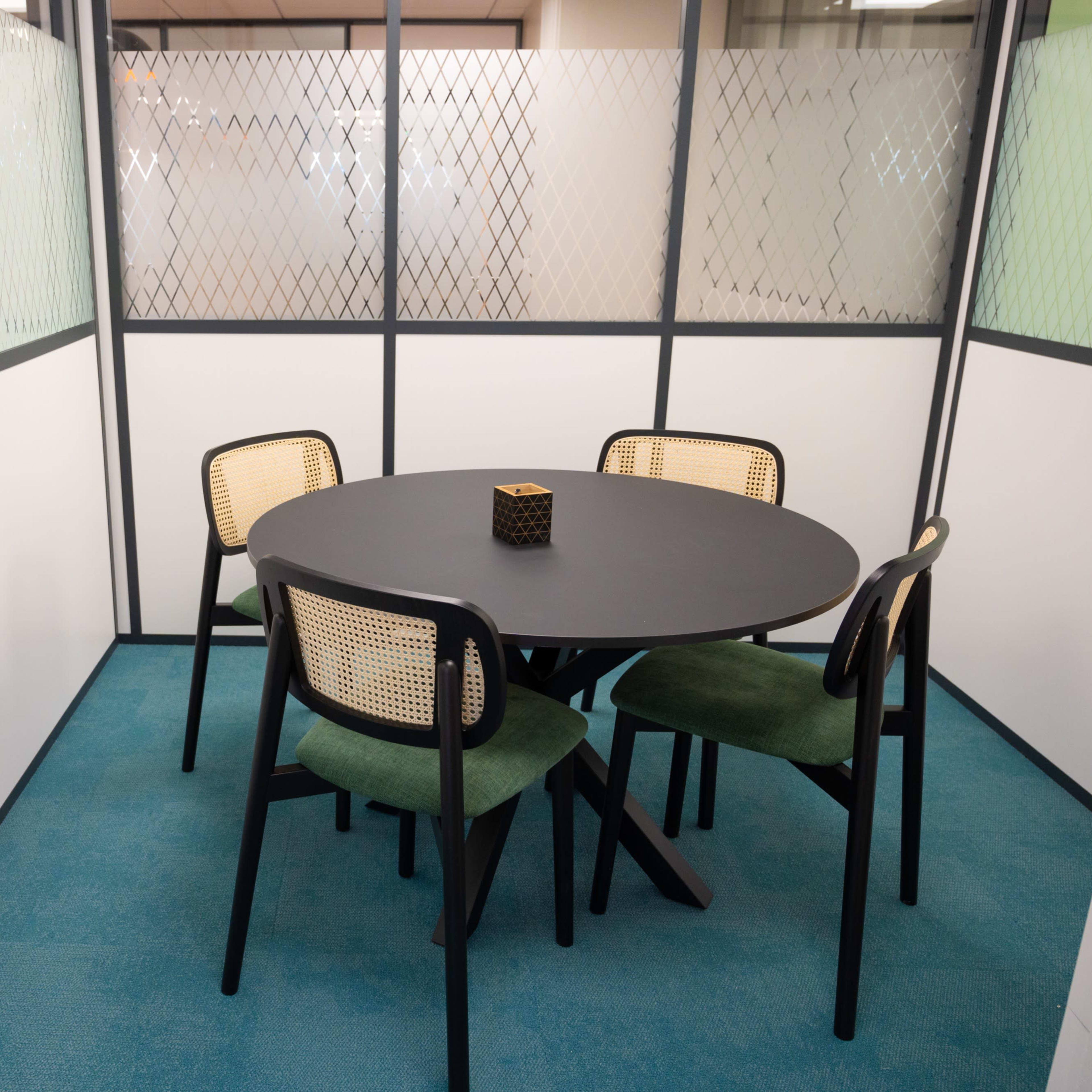 A compact meeting room features a round black table surrounded by four chairs, enclosed by glass partitions with a diamond pattern.