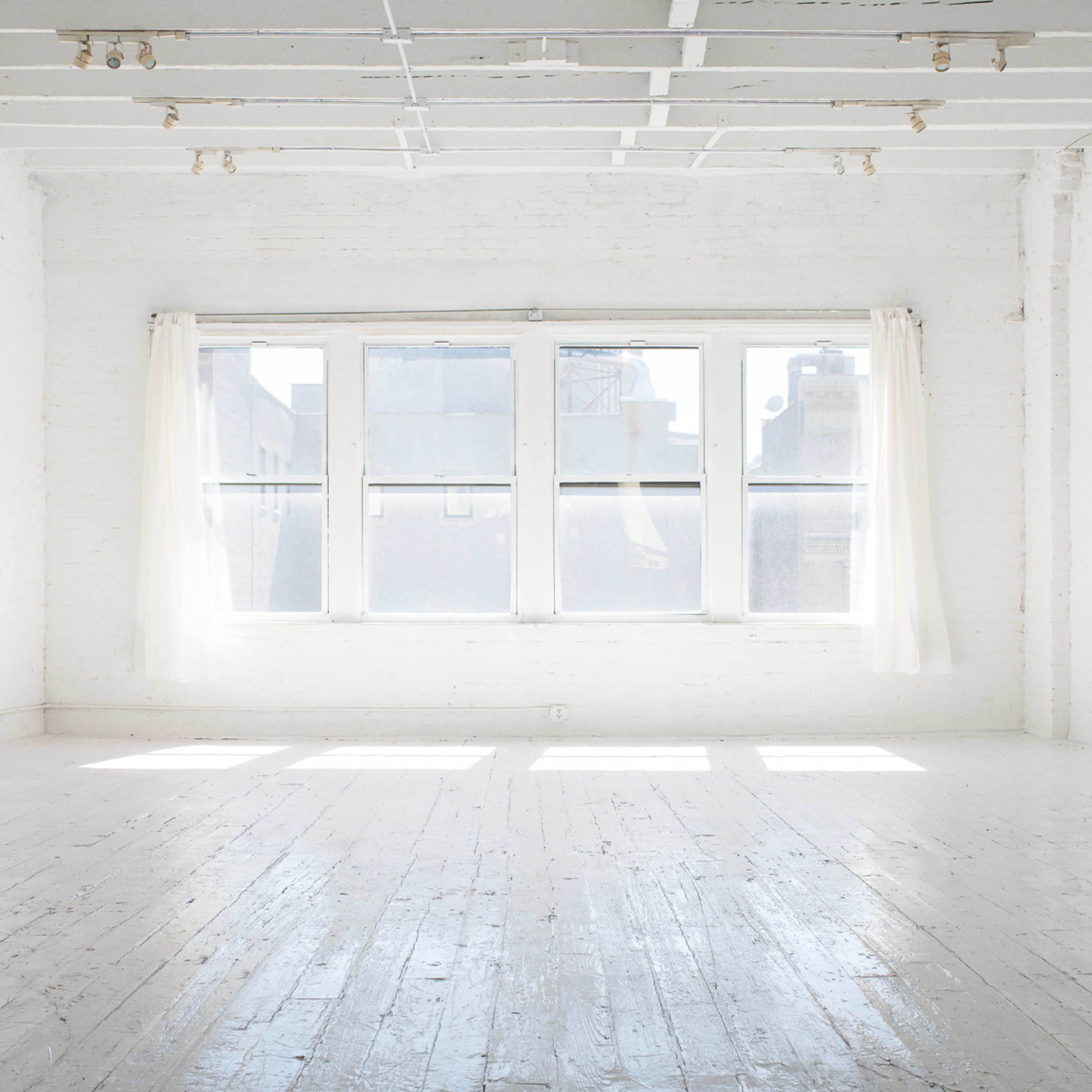 A bright, empty room with white walls and wooden flooring, featuring large windows with sheer curtains that allow light to fill the space.