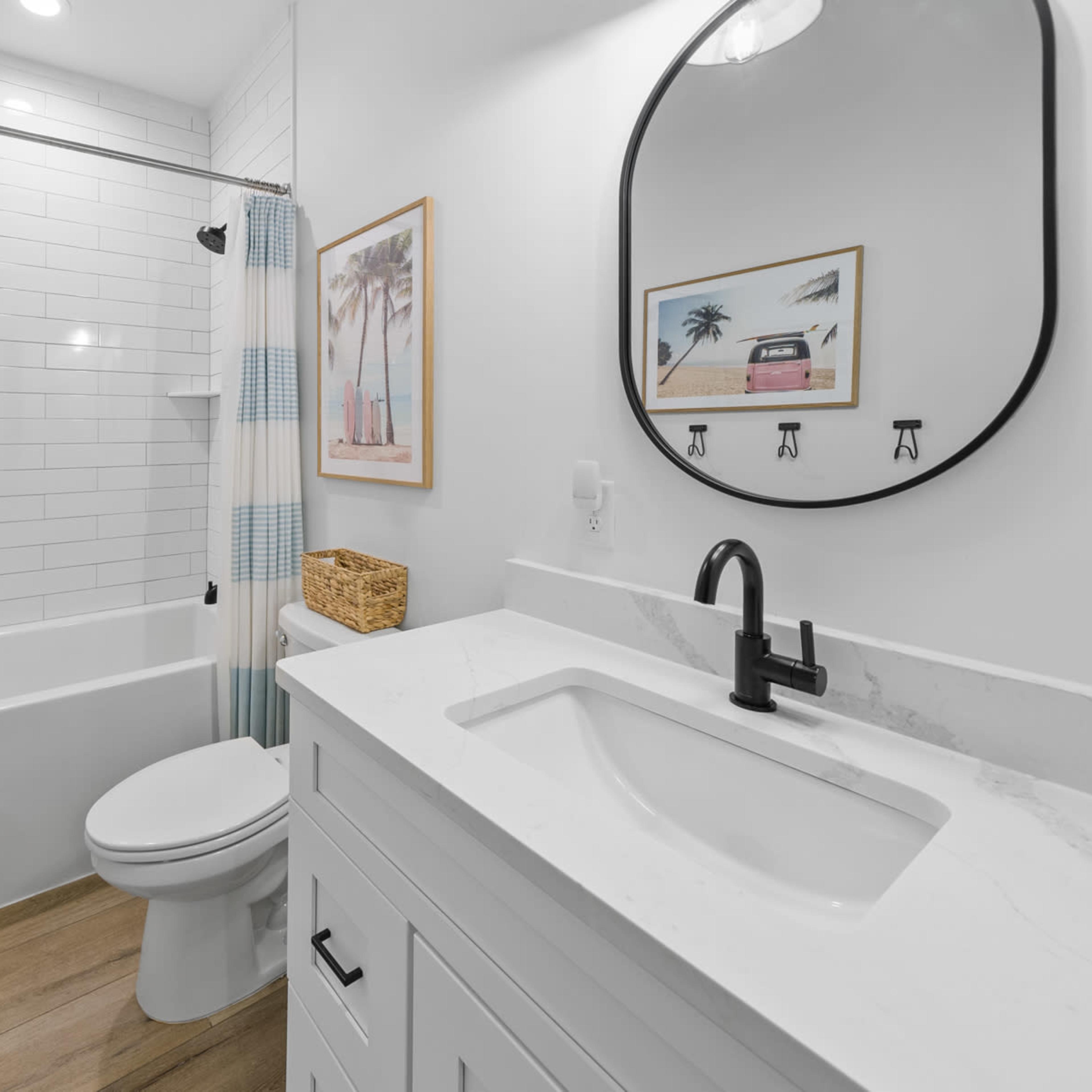 A modern bathroom featuring a white tiled bathtub with a glass shower curtain, a wall-mounted mirror, a white vanity with a sink, and wooden flooring.