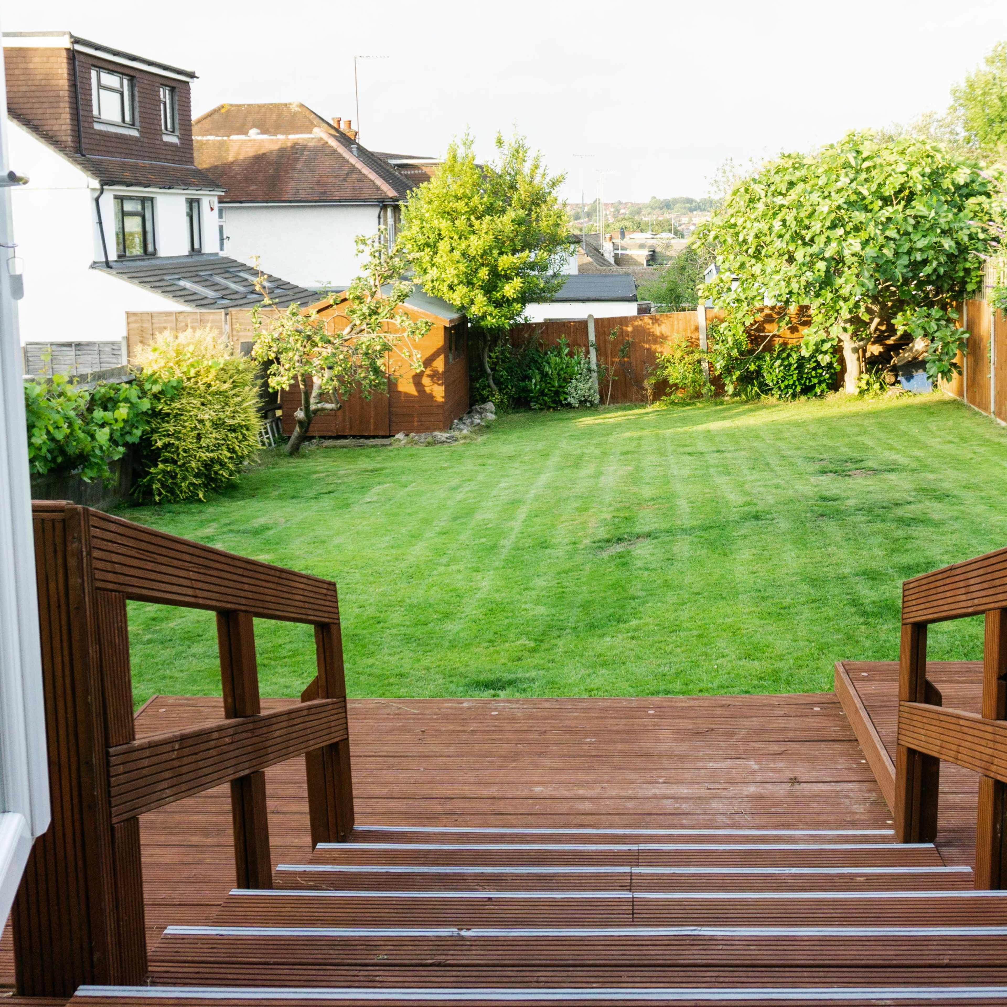 A backyard with a neatly mowed lawn, framed by wooden fences and trees, viewed from a set of wooden steps leading down from a house.