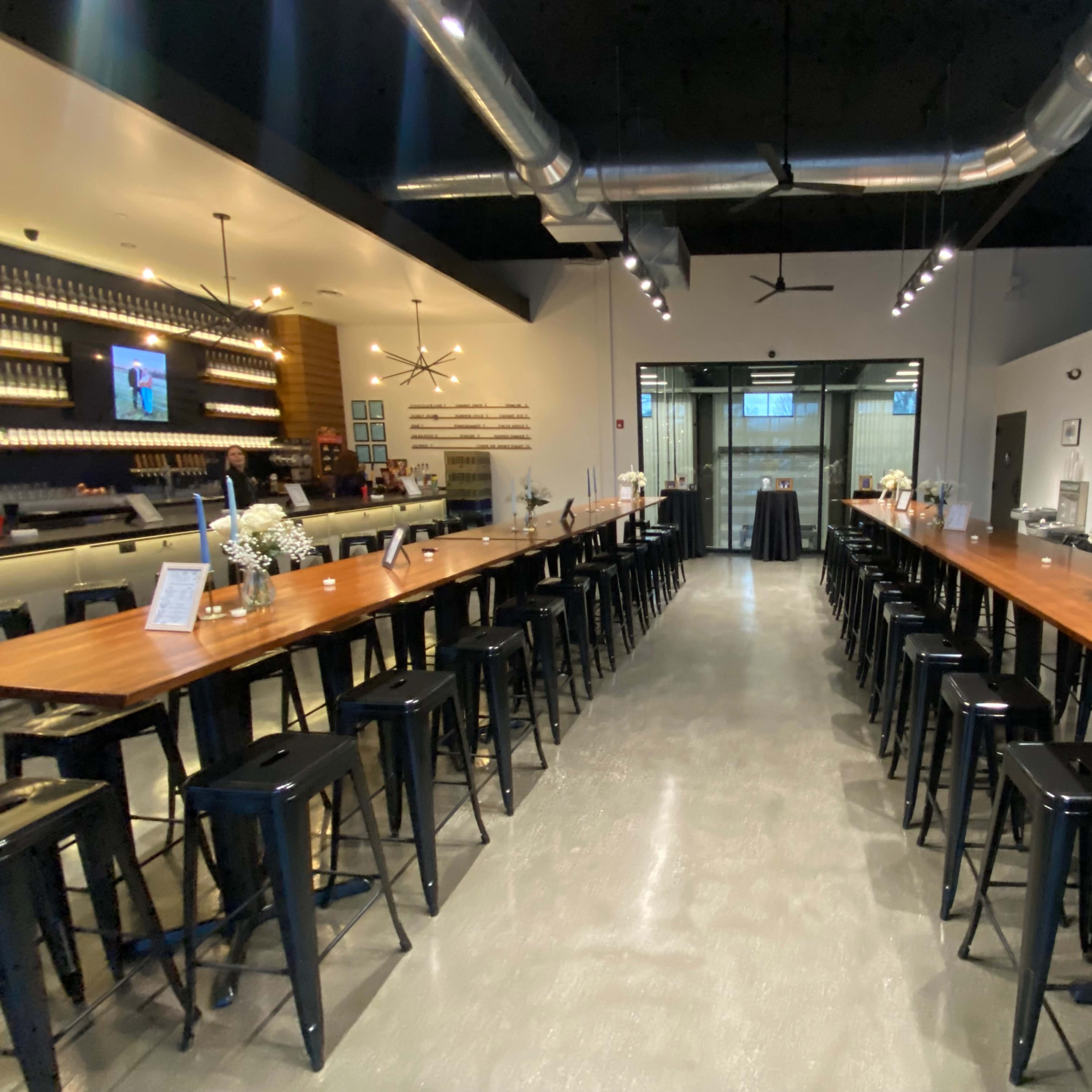 A modern bar with a long wooden counter lined with tall black stools, and shelves displaying bottles in the background.