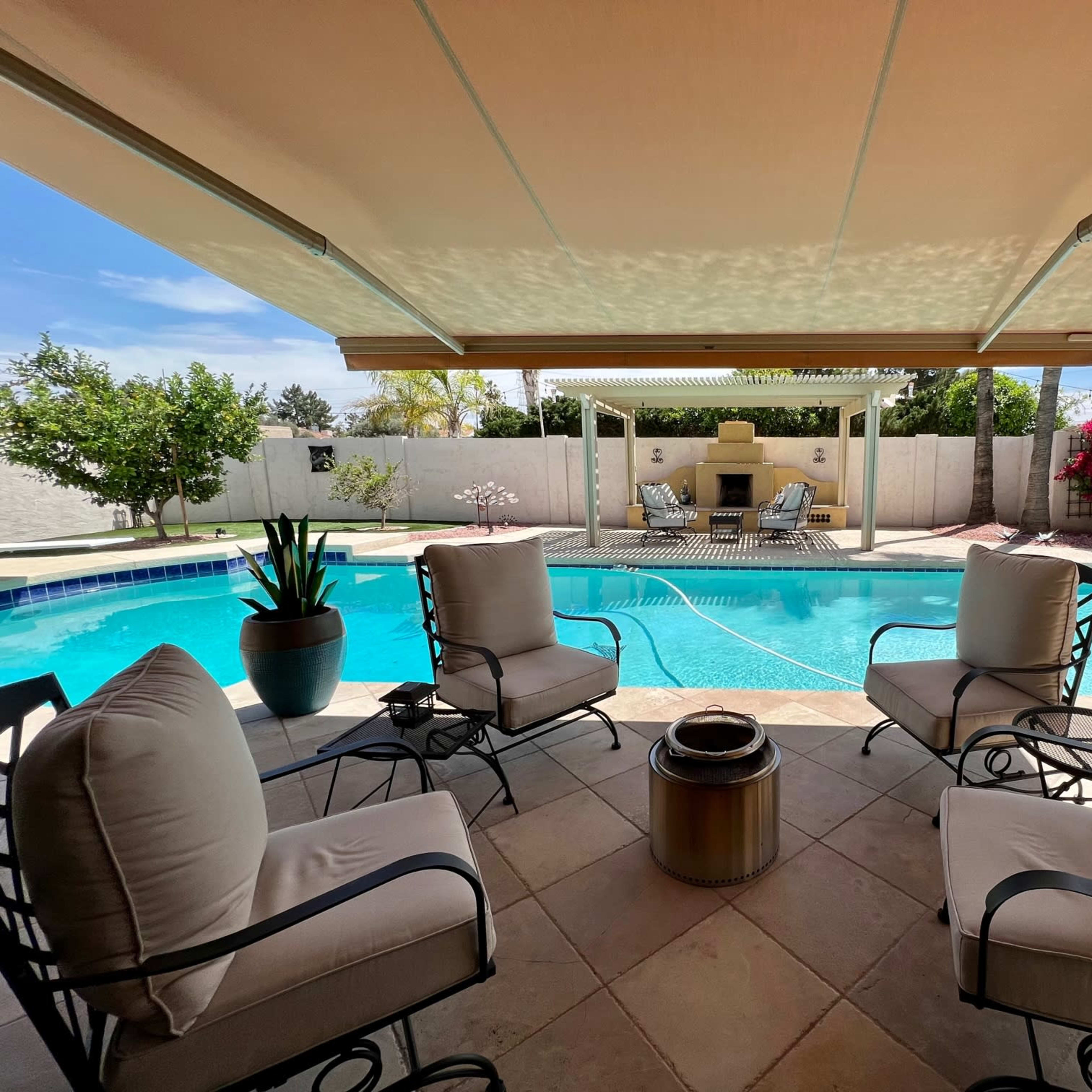 A shaded seating area with four chairs surrounds a swimming pool, framed by blooming plants and a patio enclosure.