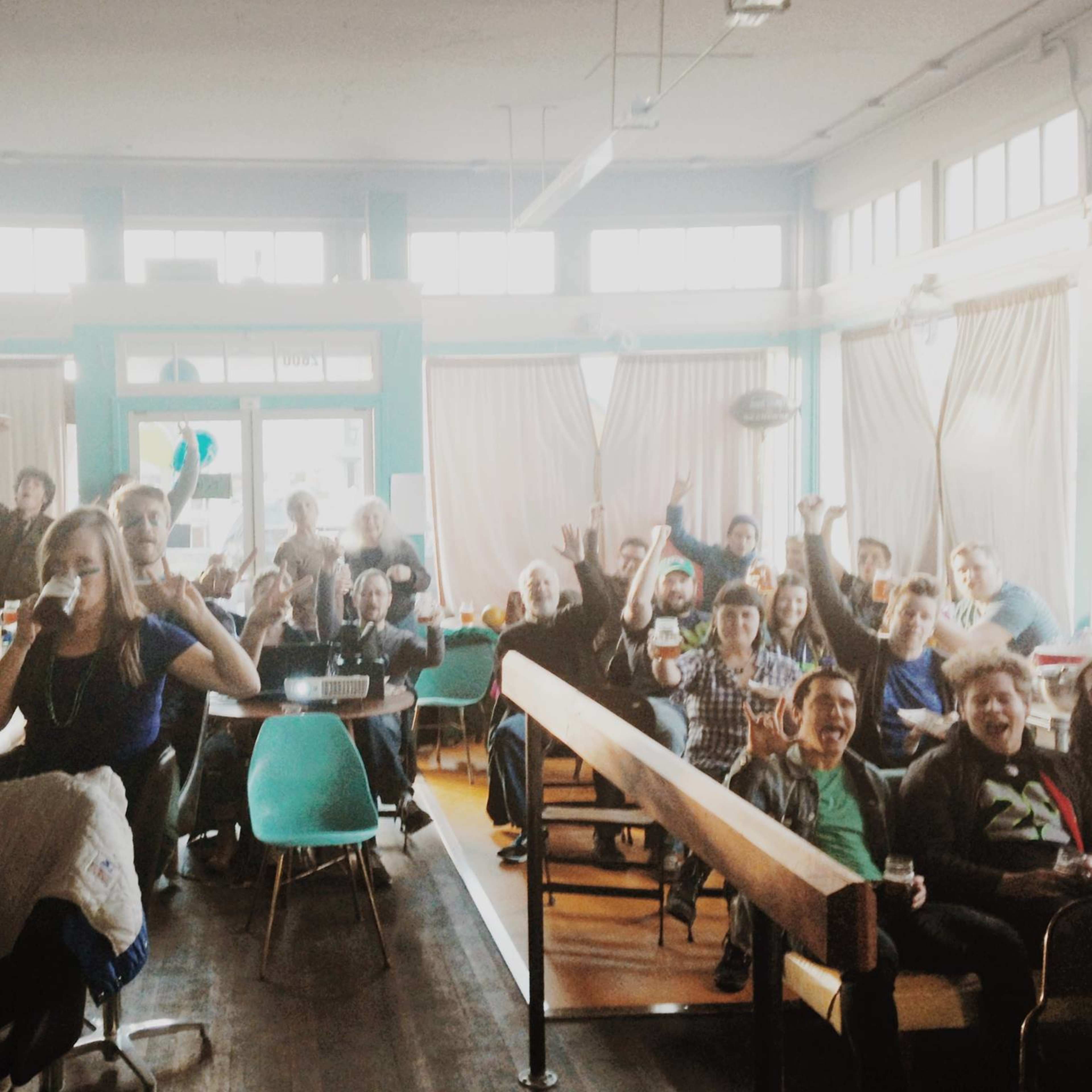 A large group of people is gathered in a lively cafe, smiling and cheering towards the camera.