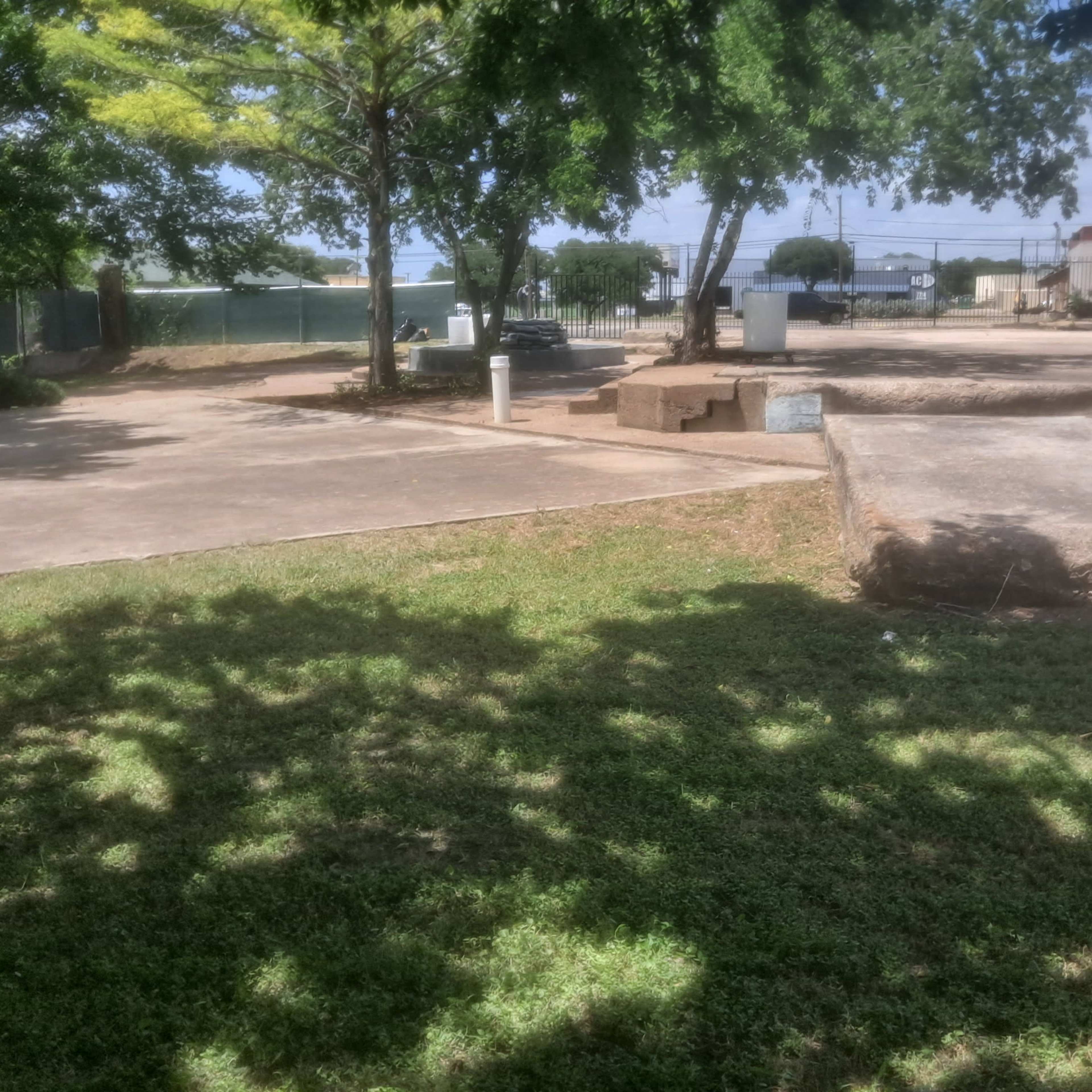 A flat, grassy area with concrete surfaces and trees in a semi-urban setting.
