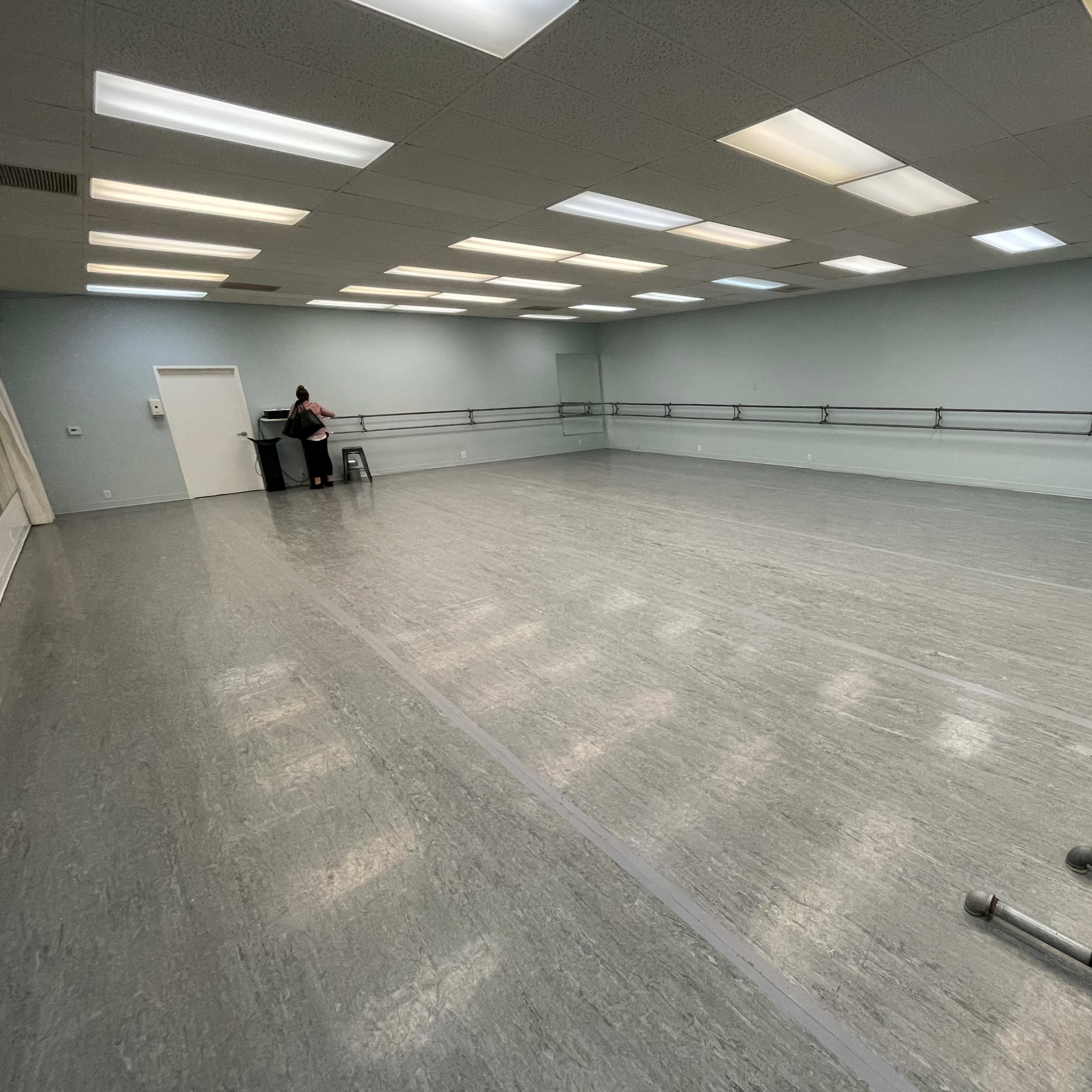 A spacious dance studio with mirrored walls, a wooden floor, and a single person sitting on a stool in the corner.