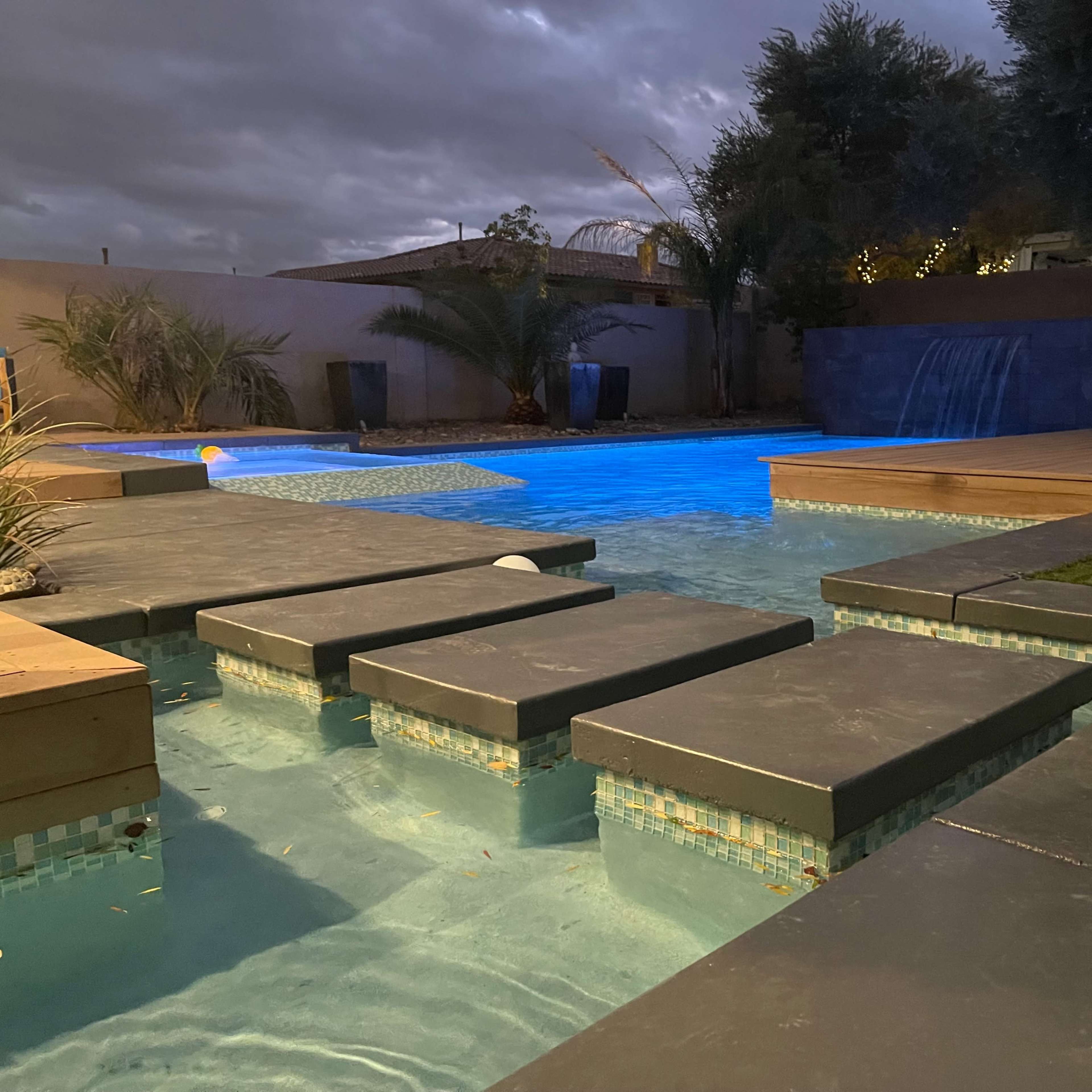 A modern pool area with illuminated water, stone pathways, and a waterfall feature in the background.