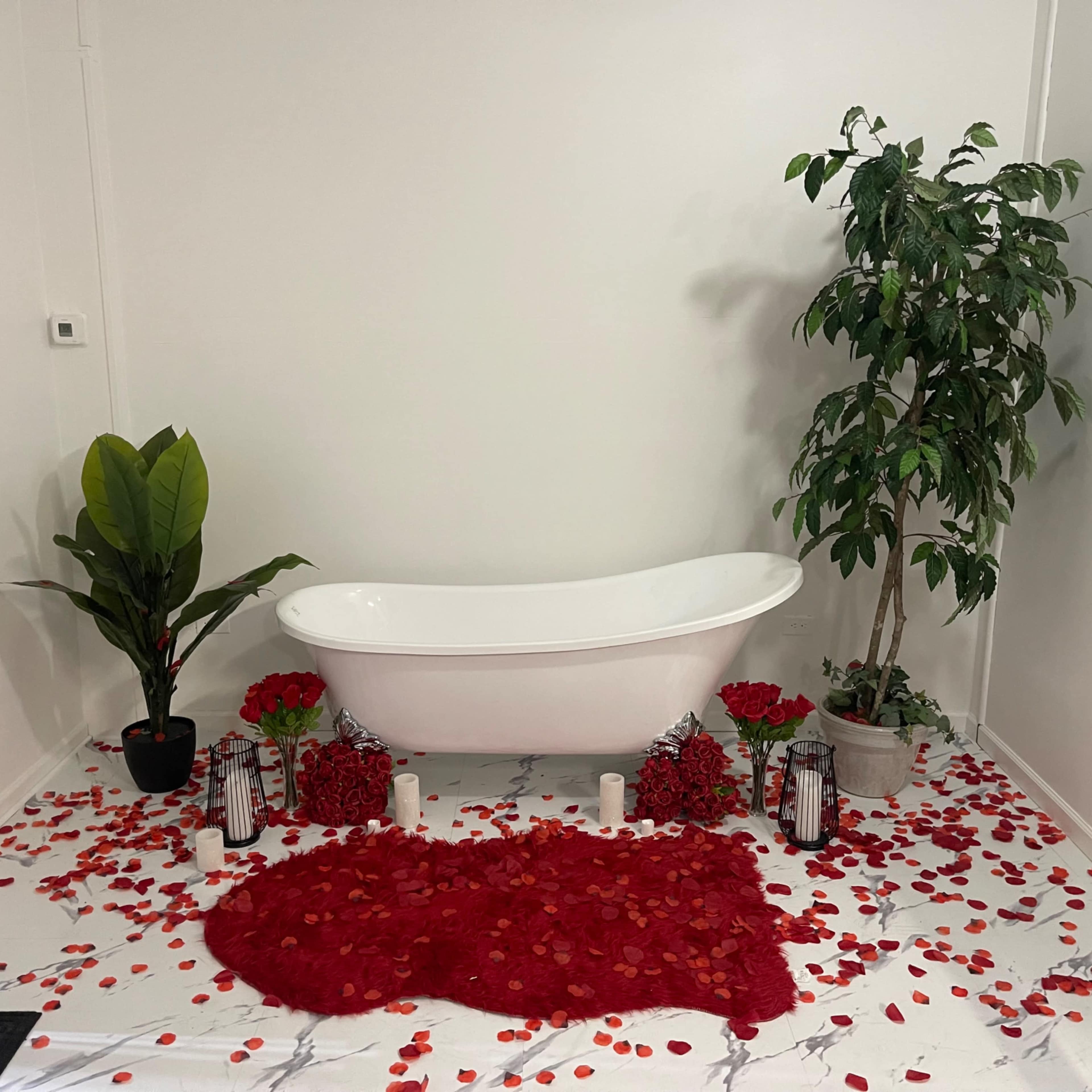 A white freestanding bathtub is surrounded by red rose petals, candles, and decorative plants in a minimalistic bathroom setting.