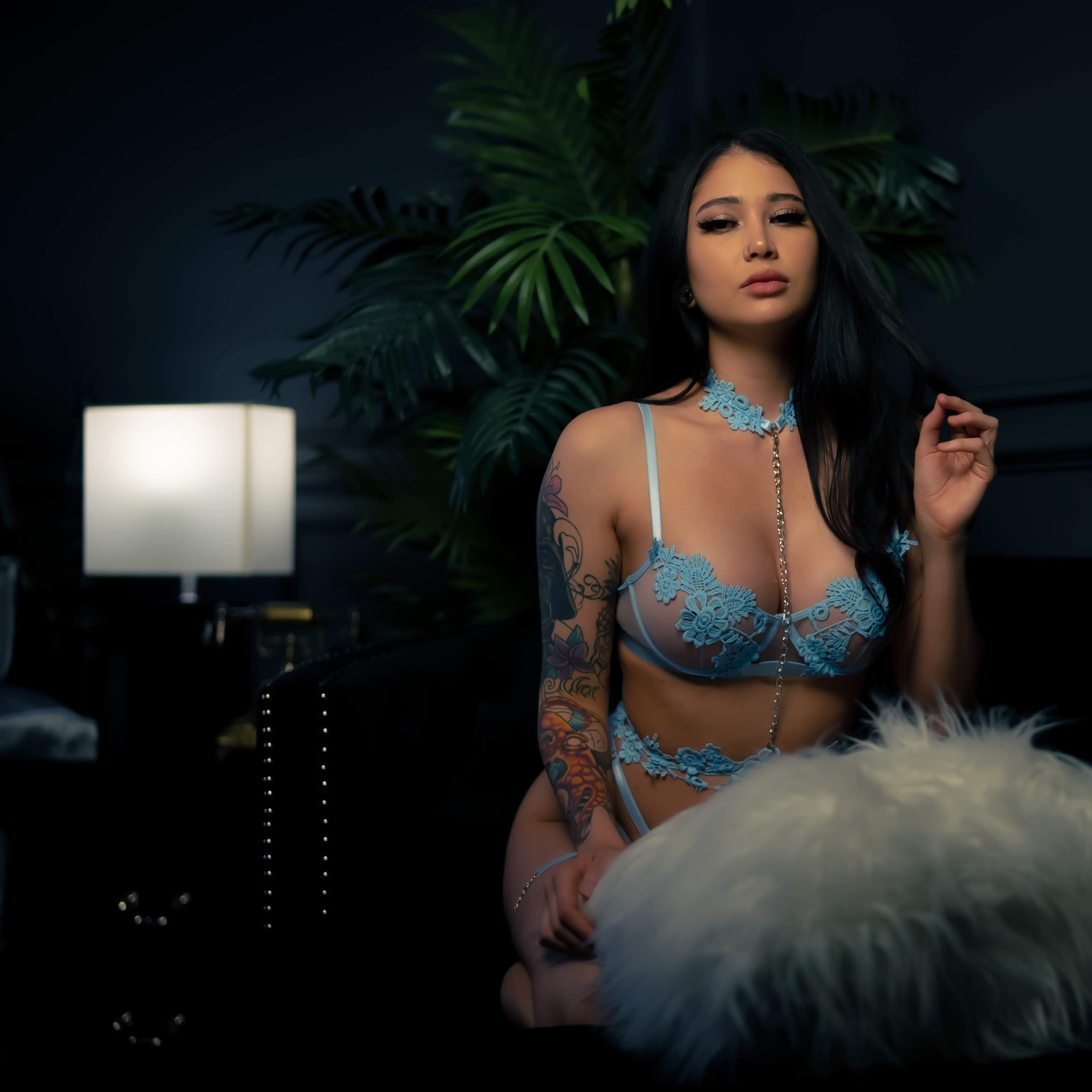 A woman with long dark hair and tattoos poses on a dark couch surrounded by plants and soft lighting.
