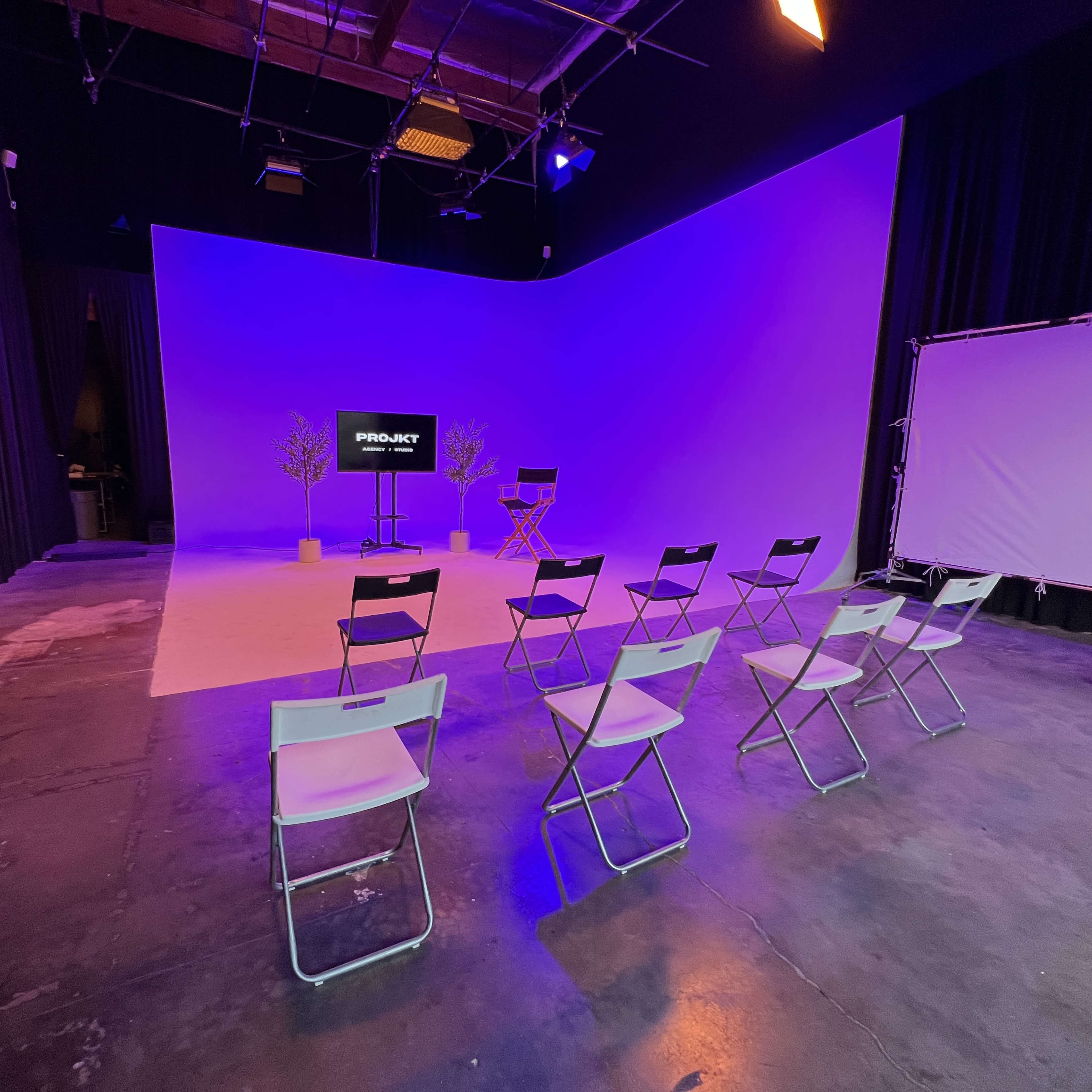A studio setup features a row of white folding chairs facing a screen displaying "PRO-ART," with purple-backlit walls and two small trees in the background.