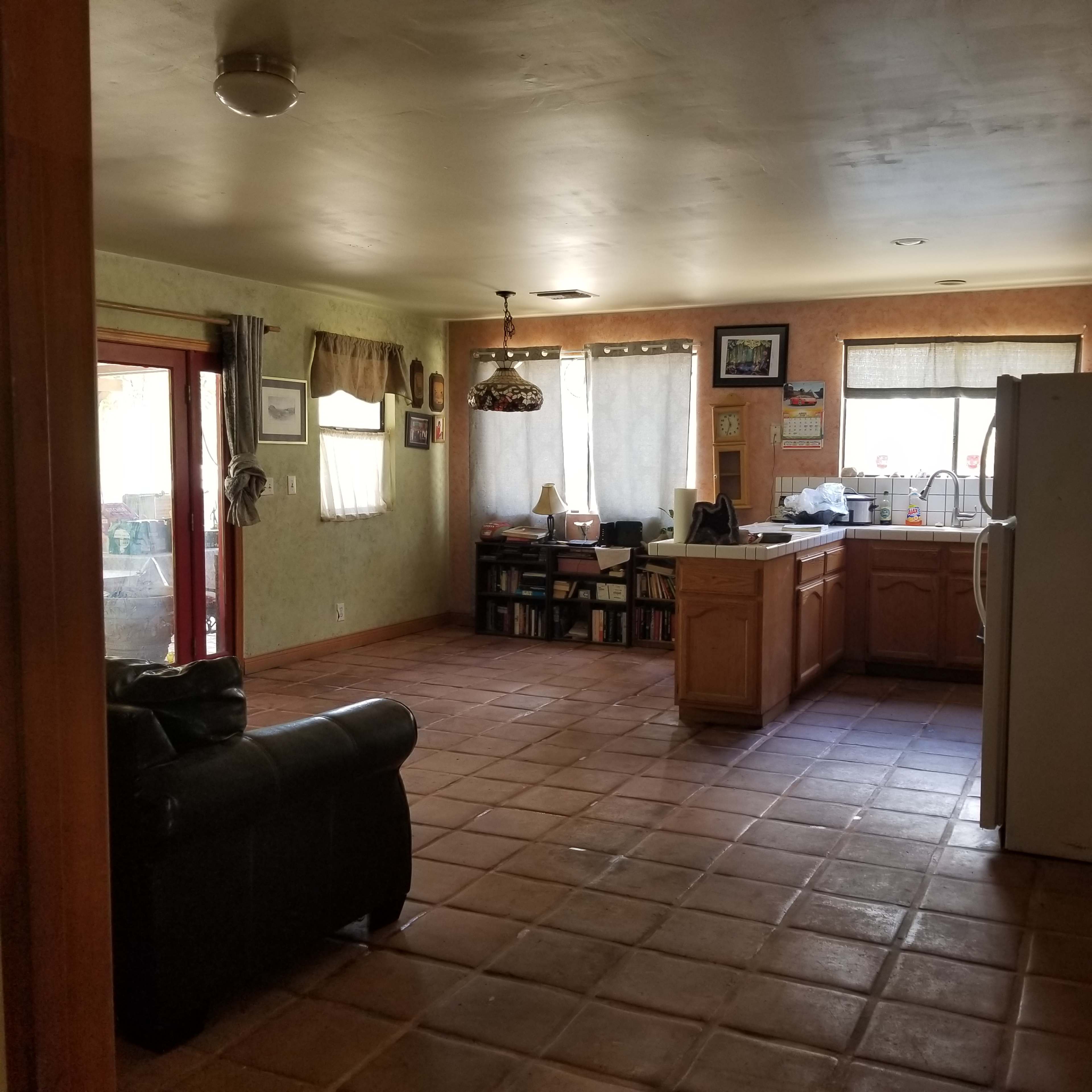 A spacious kitchen features a tiled floor, wooden cabinets, and an adjacent living area with a black couch and large windows.