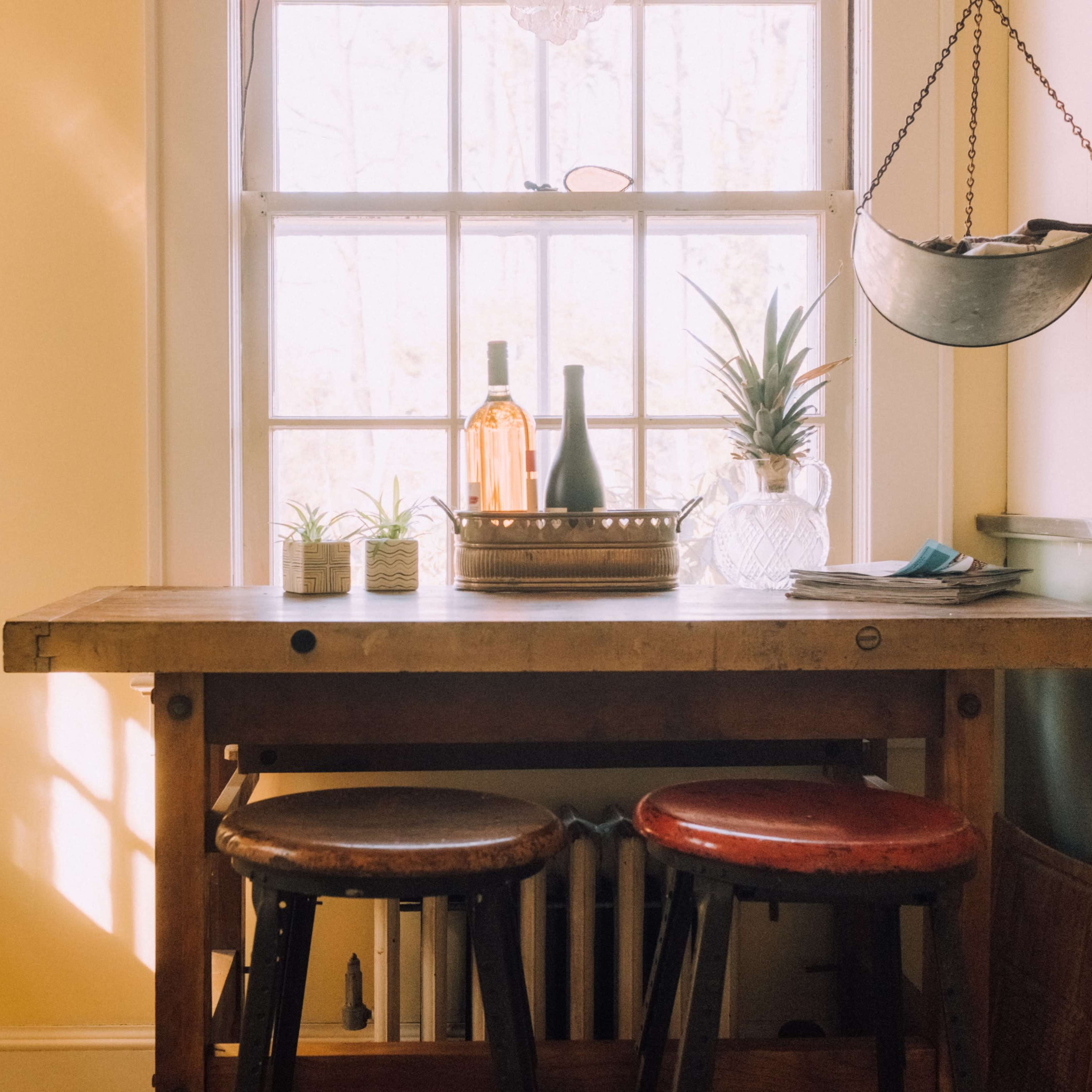 A wooden table with two stools sits by a window, displaying bottles and plants on a sunny afternoon.