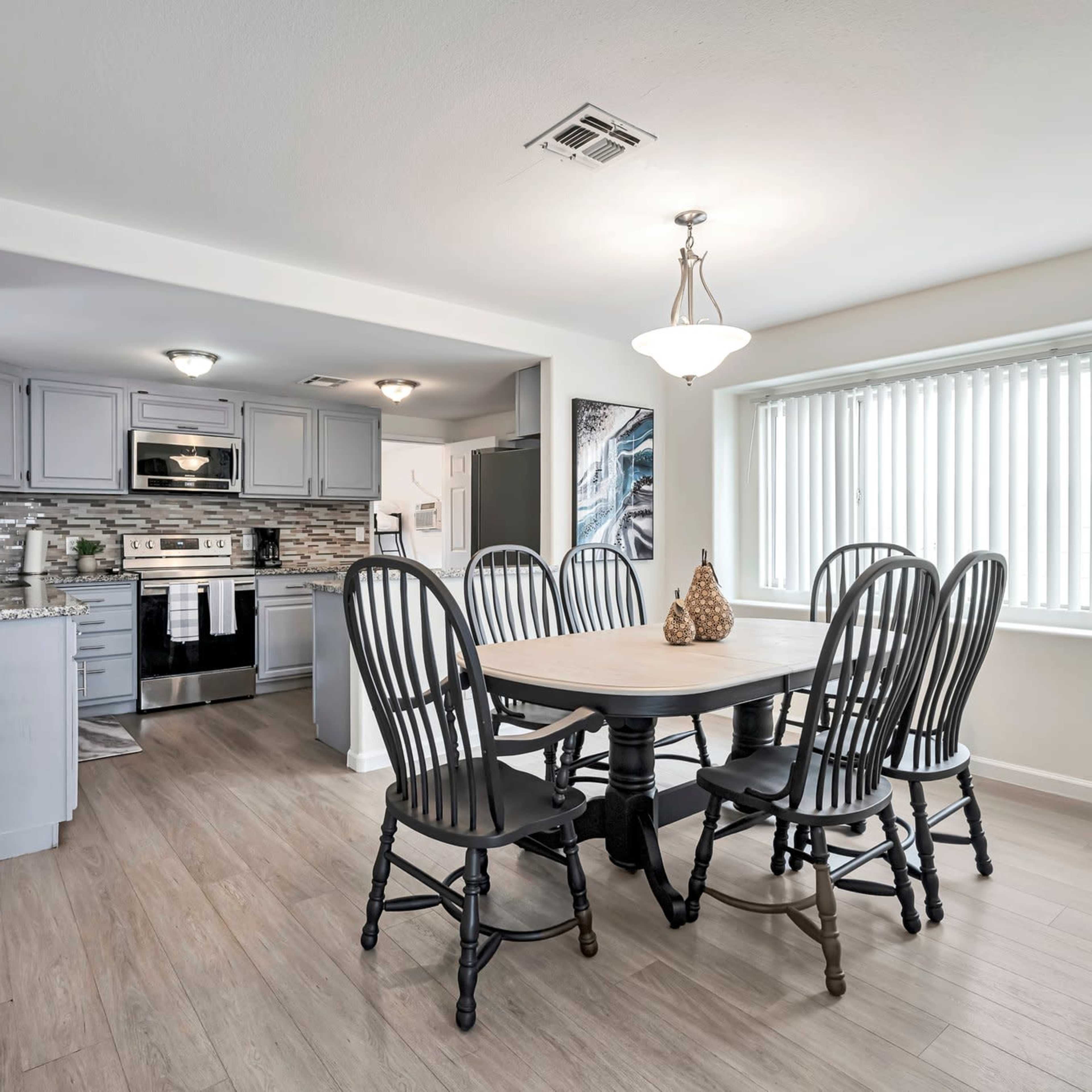 A dining area features a wooden table surrounded by six black chairs, with a kitchen visible in the background.
