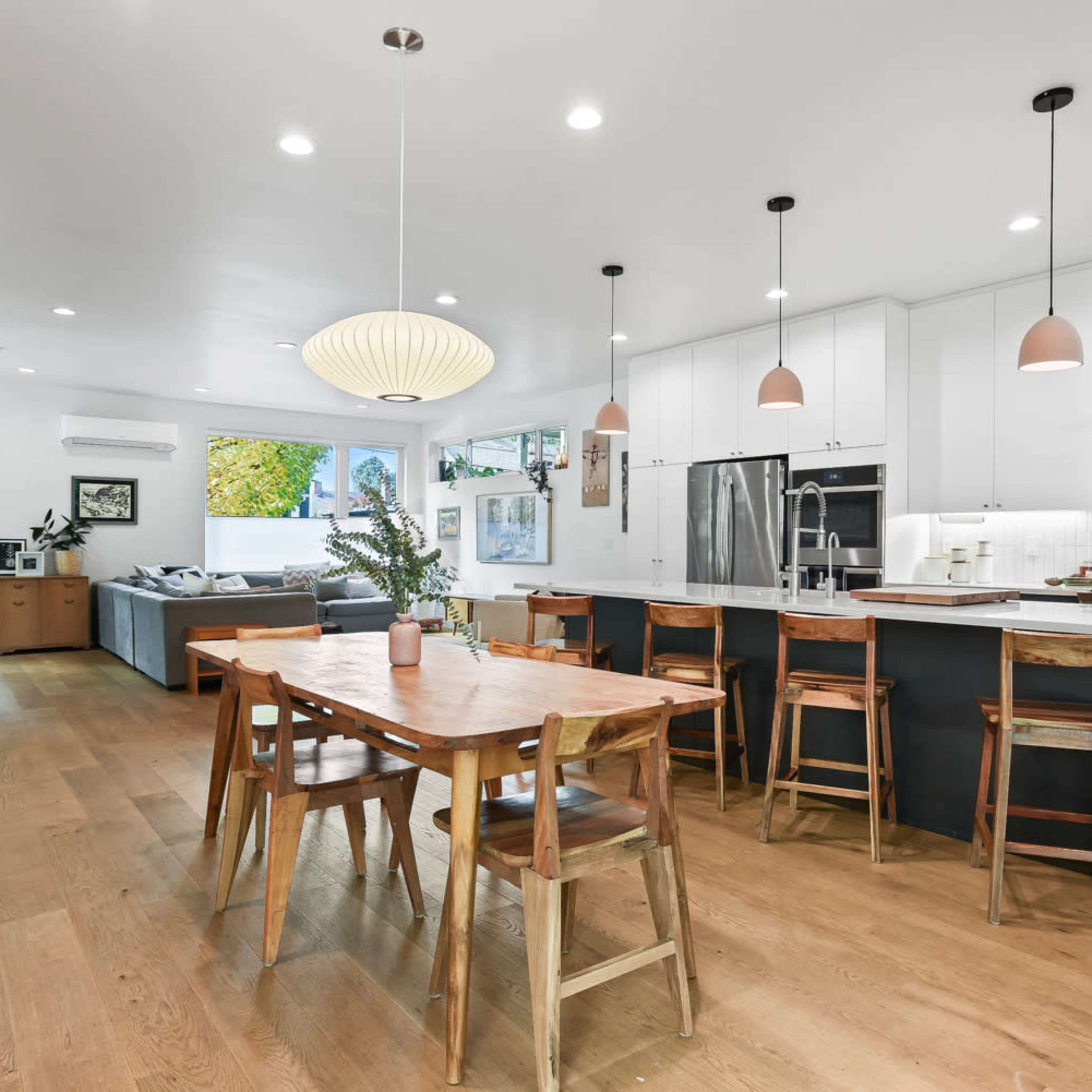 A modern kitchen and dining area features a wooden table surrounded by chairs, sleek cabinetry, and large windows allowing natural light to fill the space.